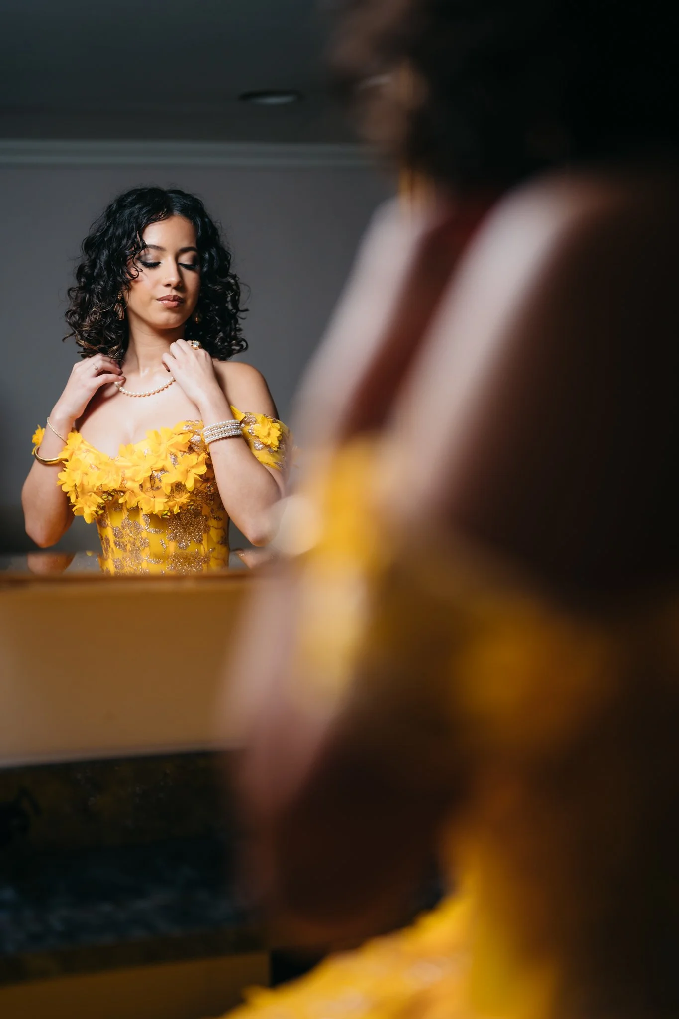 Woman in a yellow dress with floral details and jewelry looking at herself in a mirror.