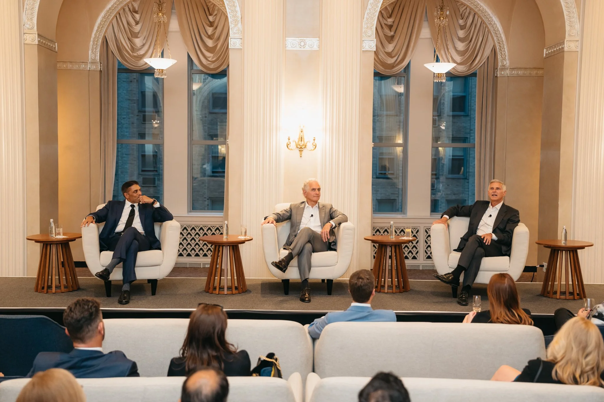 Three men seated on stage in armchairs during a panel discussion or conference, with an audience watching. The background features tall windows with drapes and elegant decor.