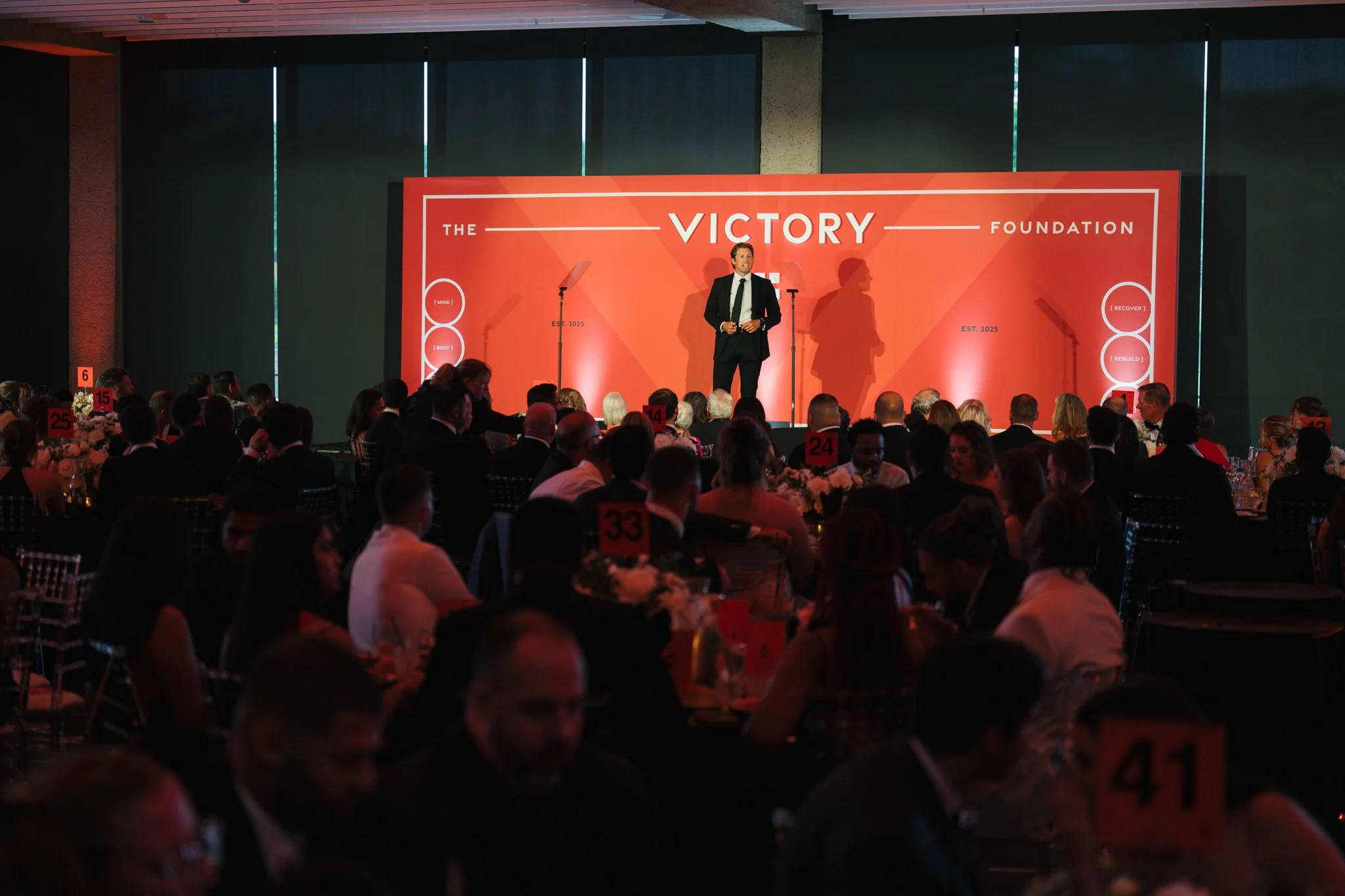 A speaker in a black suit standing on stage at a formal event with a large red backdrop that reads "The Victory Foundation." The audience seated at tables watching the speaker. The event appears to be in a spacious venue with high ceilings.