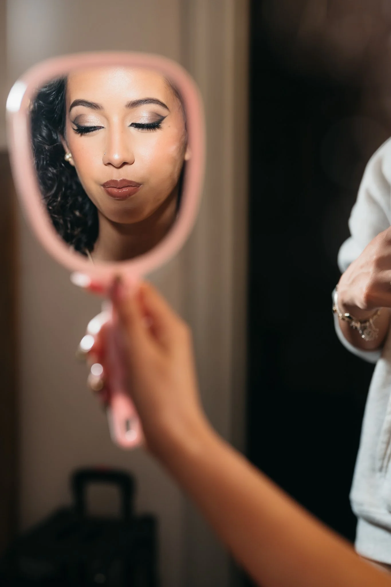 A woman with makeup and earrings looks at her reflection in a pink handheld mirror, which is held by another person.