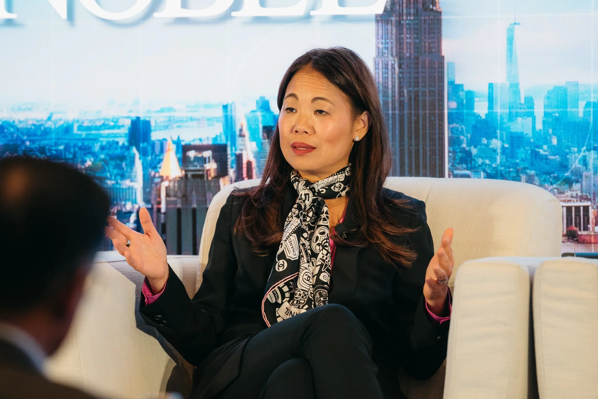 A woman with medium-length dark hair and wearing a black blazer and patterned scarf, sitting on a beige sofa, gesturing with her hands, during a discussion or interview against a backdrop of a city skyline.