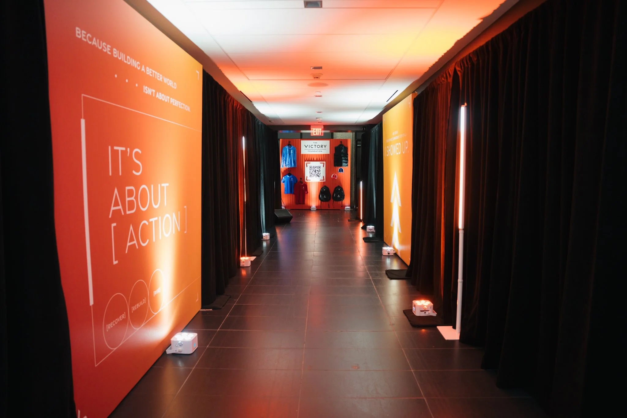 A hallway with black curtains on both sides, illuminated by orange vertical lights. Large orange banners with motivational quotes are displayed on the walls. At the end of the hallway, there is a red wall with clothing items and a QR code, and a sign