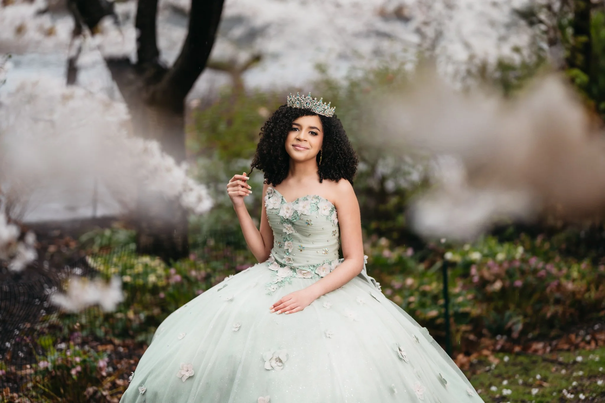 A young woman in a light green ball gown with floral embellishments, wearing a crown, sitting outdoors among flowering trees.