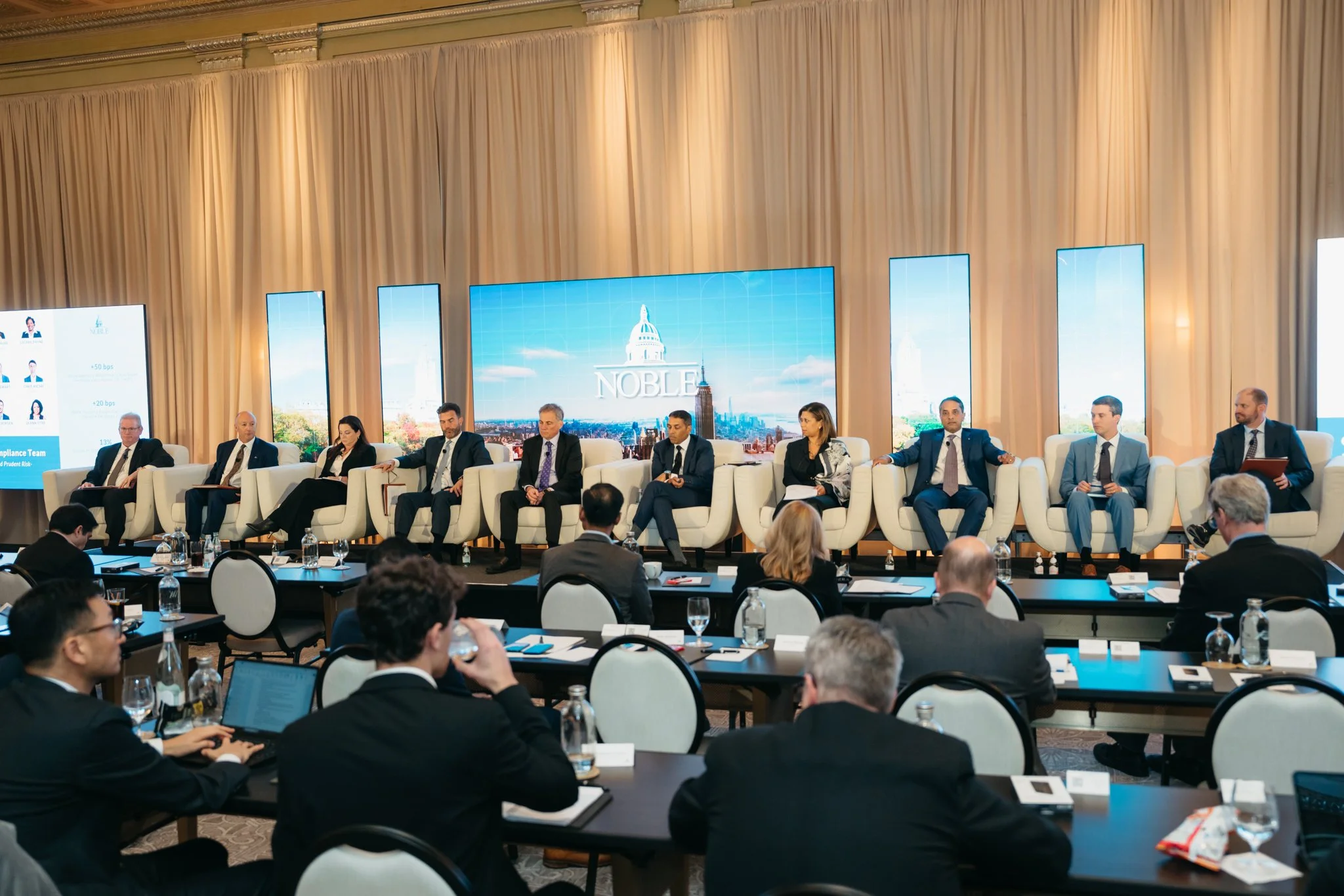Panel of ten professionals seated on stage at a conference, with a large screen displaying the word 'NOBLE' behind them, in a room with beige curtains and audience members taking notes.