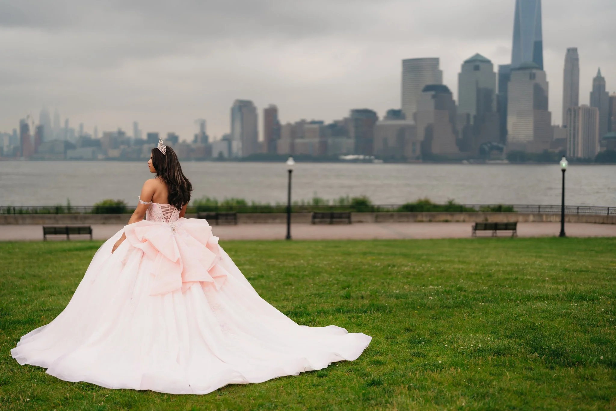 A woman wearing a pink ball gown and tiara standing on a grassy area near a river in front of a city skyline with tall buildings, overcast sky.