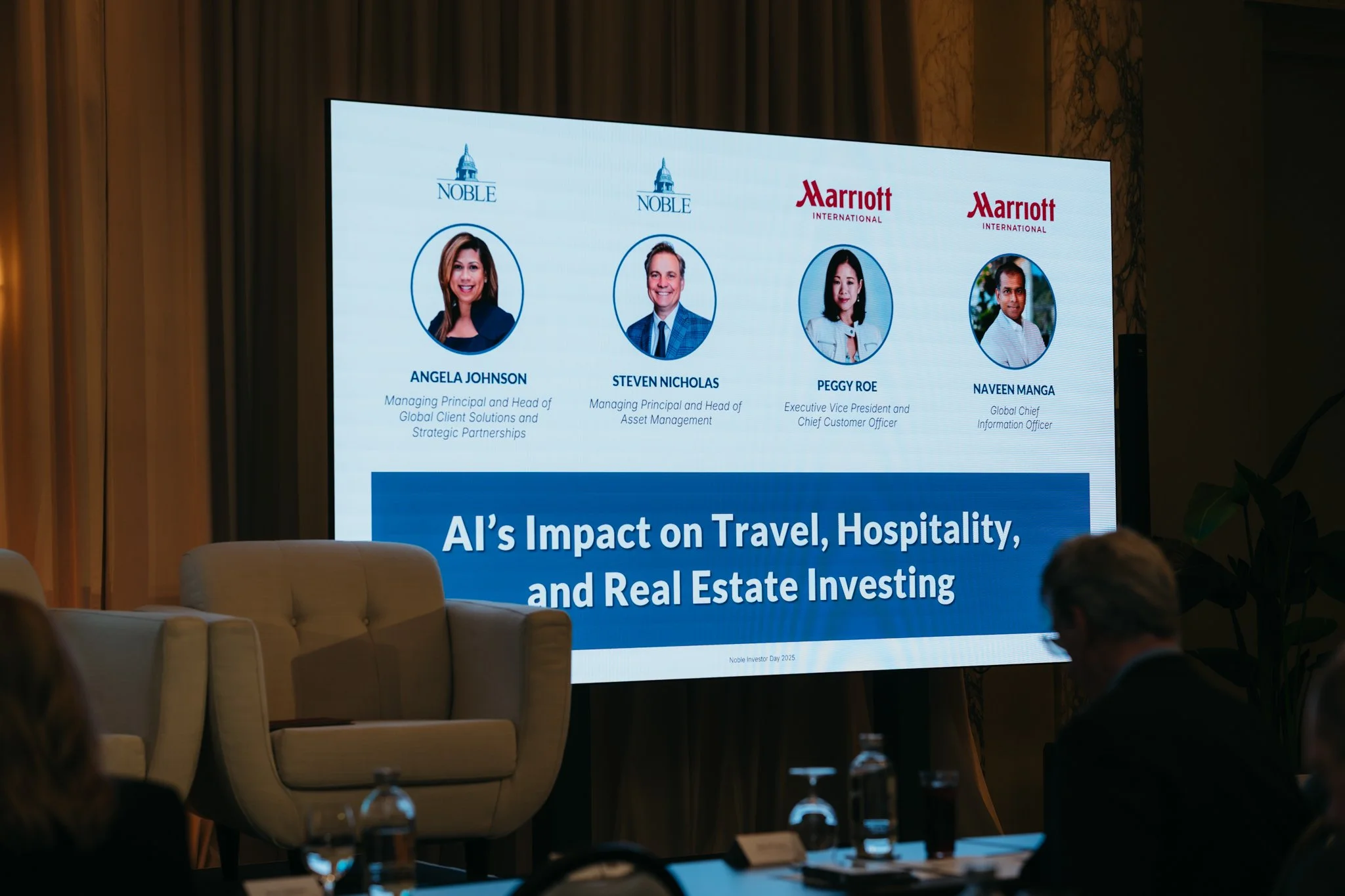 A large screen displaying the names, photos, and titles of four speakers at a conference on AI's impact on travel, hospitality, and real estate investing, with chairs and audience members visible in the foreground.
