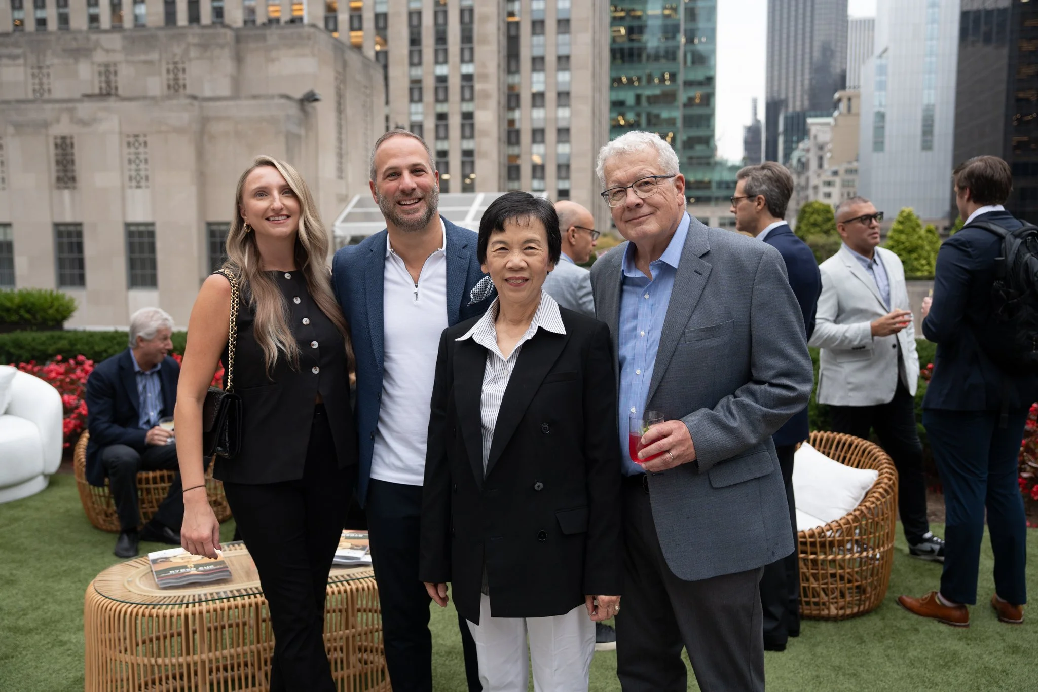 A diverse group of five people smiling and posing together at a rooftop gathering in an urban setting, with skyscrapers and city buildings in the background.