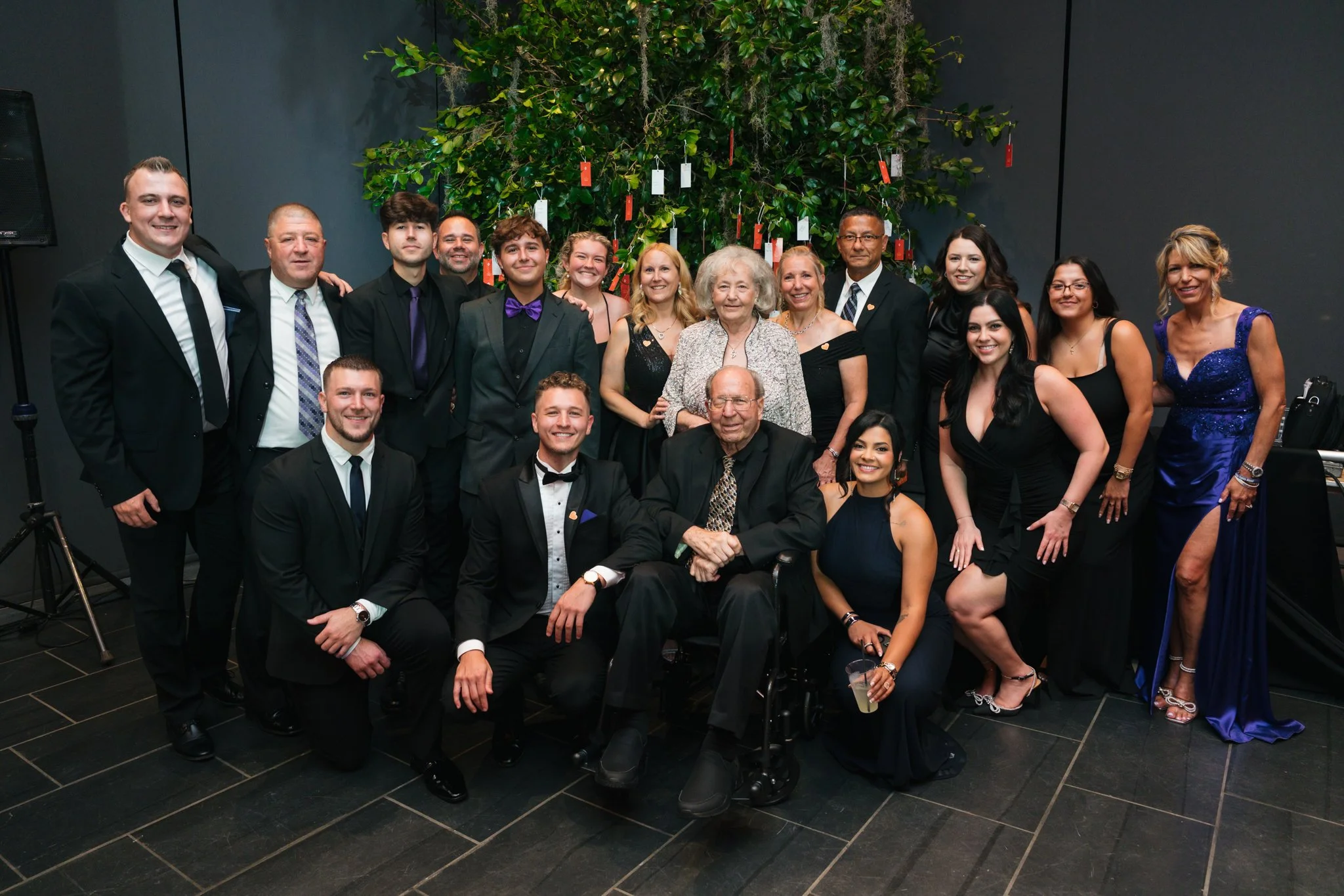 Group of people dressed in formal attire posing in front of a green tree with red and white tags hanging from it, at an indoor event.