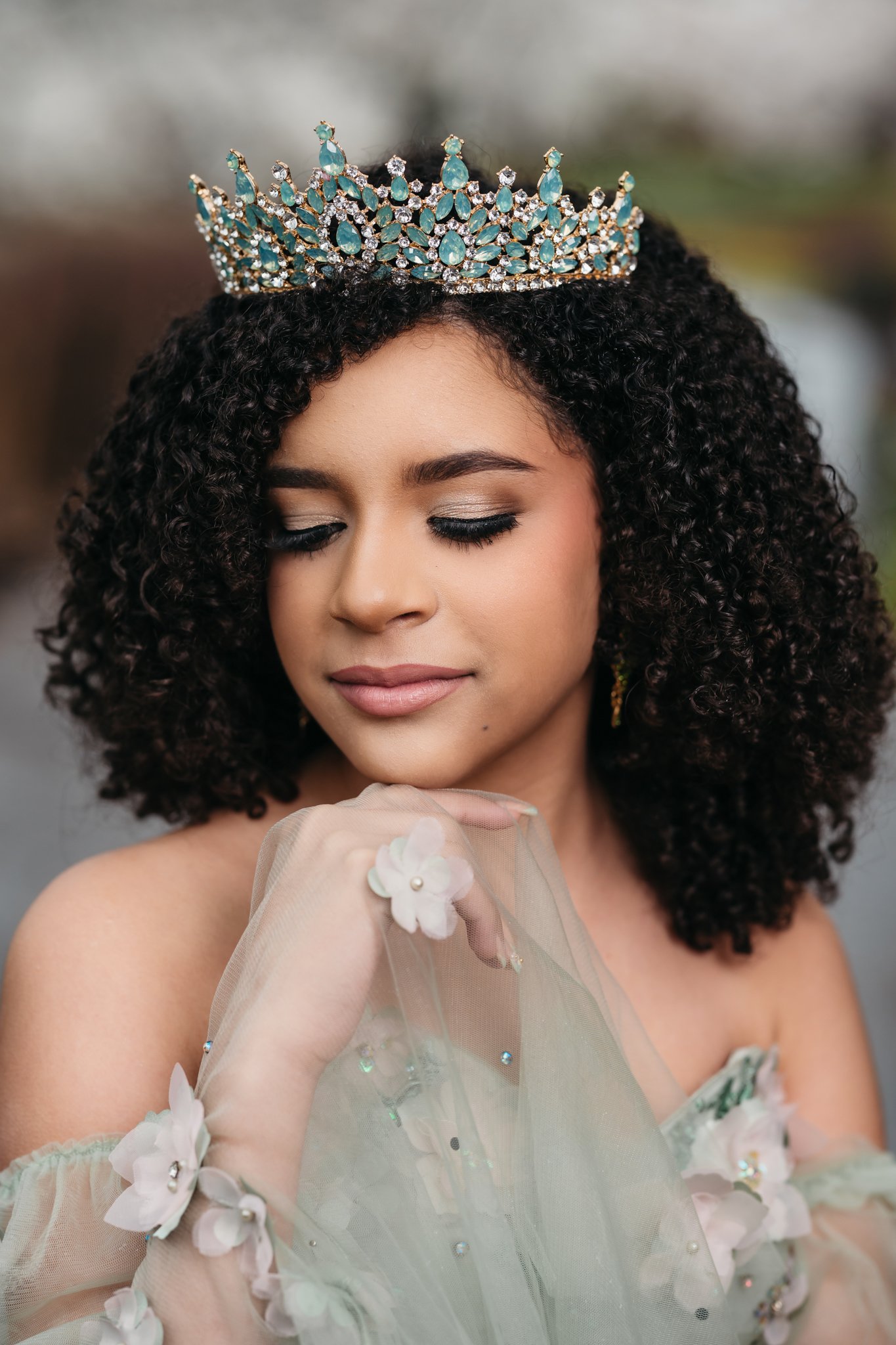 Young woman with curly hair wearing a crown and a sheer dress with floral embellishments, gently touching her face with her eyes closed.
