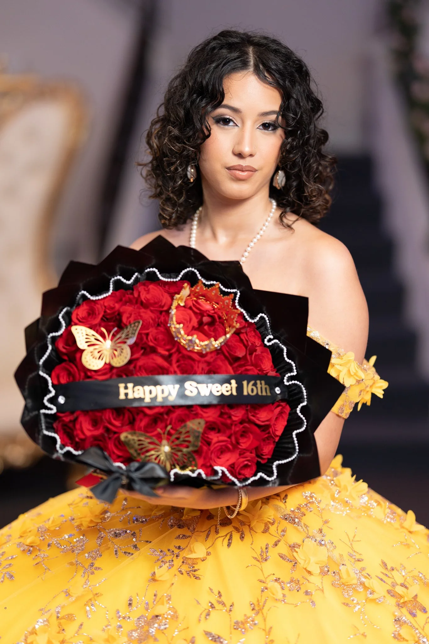 A young woman in a yellow, floral, beaded dress holding a bouquet of red roses with a 'Happy Sweet 16th' message, decorated with gold butterflies and jewelry.