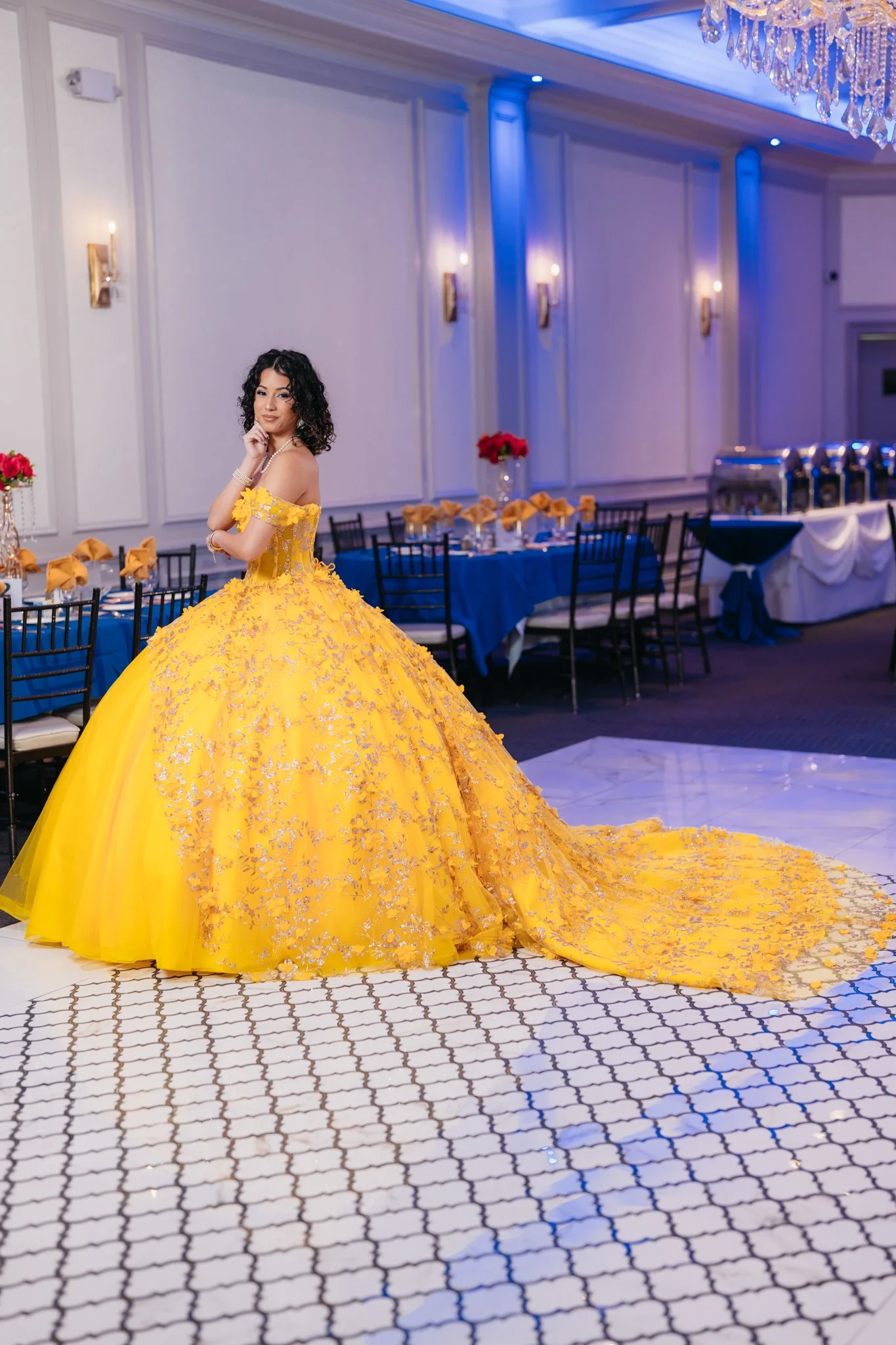 A woman in a yellow ball gown with silver embroidery stands in a decorated banquet hall.
