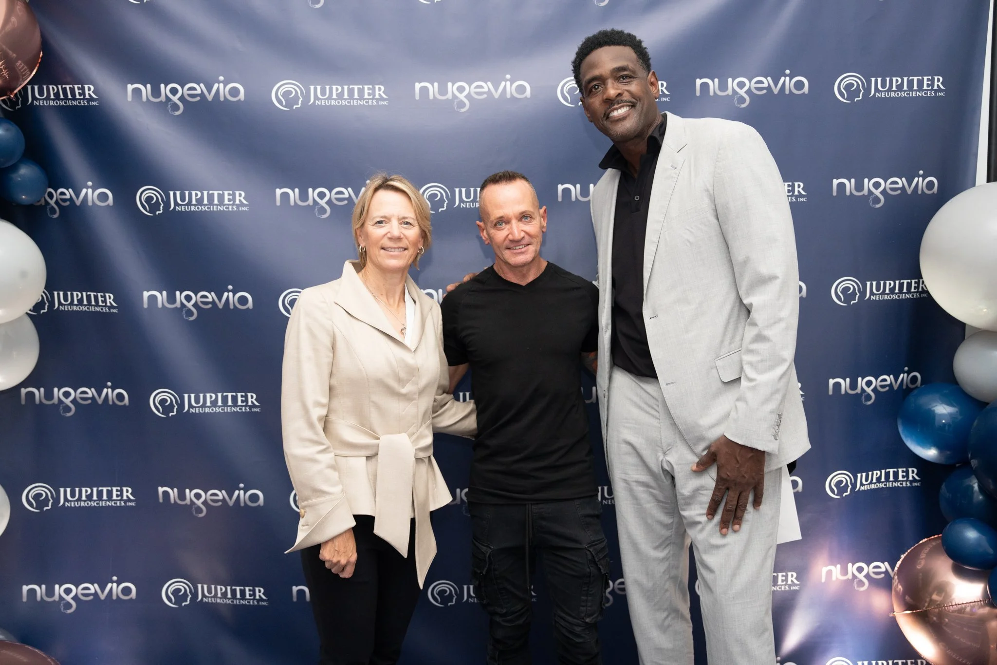 Three people posing in front of a blue backdrop with logos for Nugevia and Jupiter Neurosciences, with balloons on the sides.