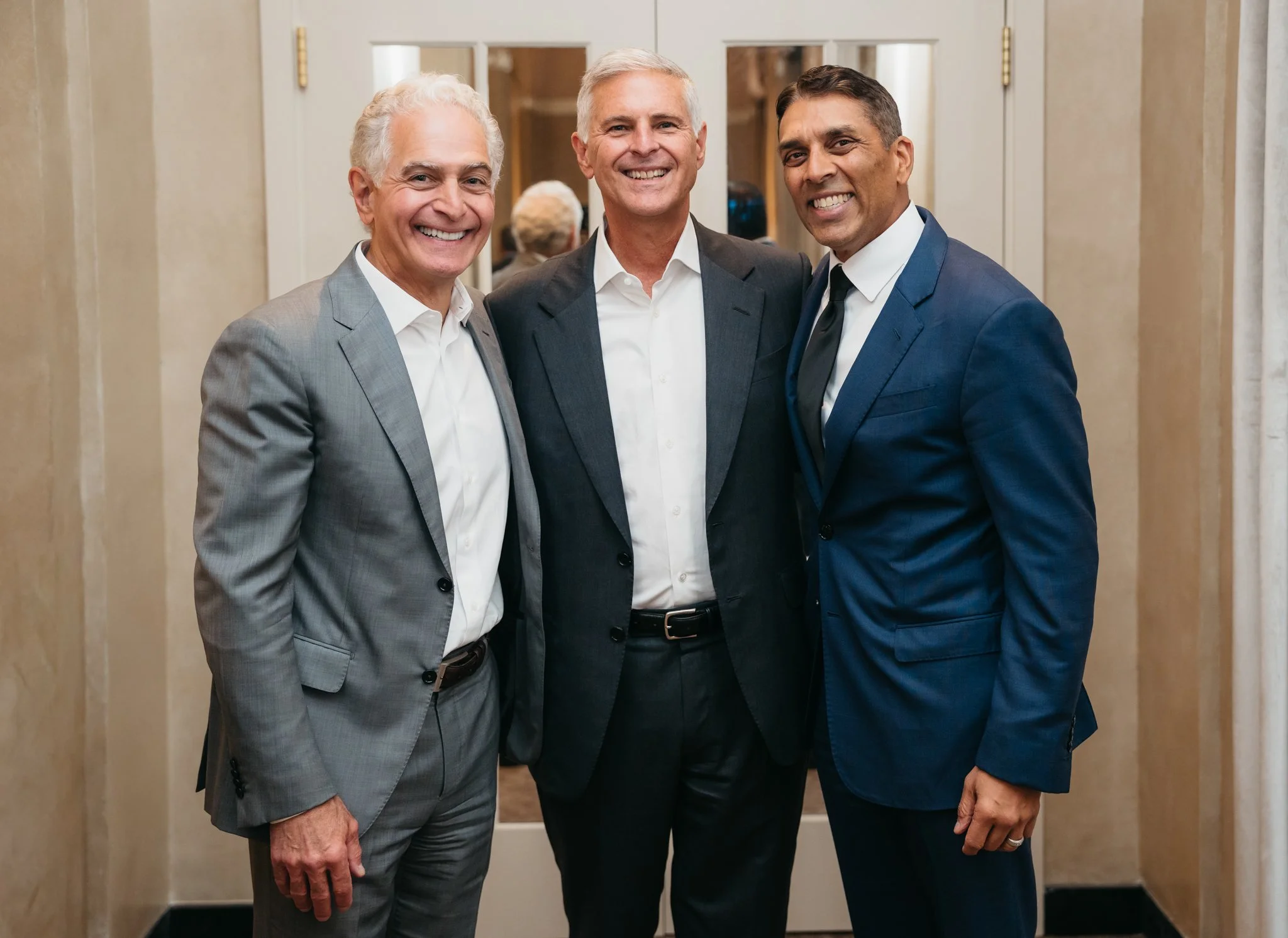 Three men in business suits smiling and standing together indoors in front of a mirror.