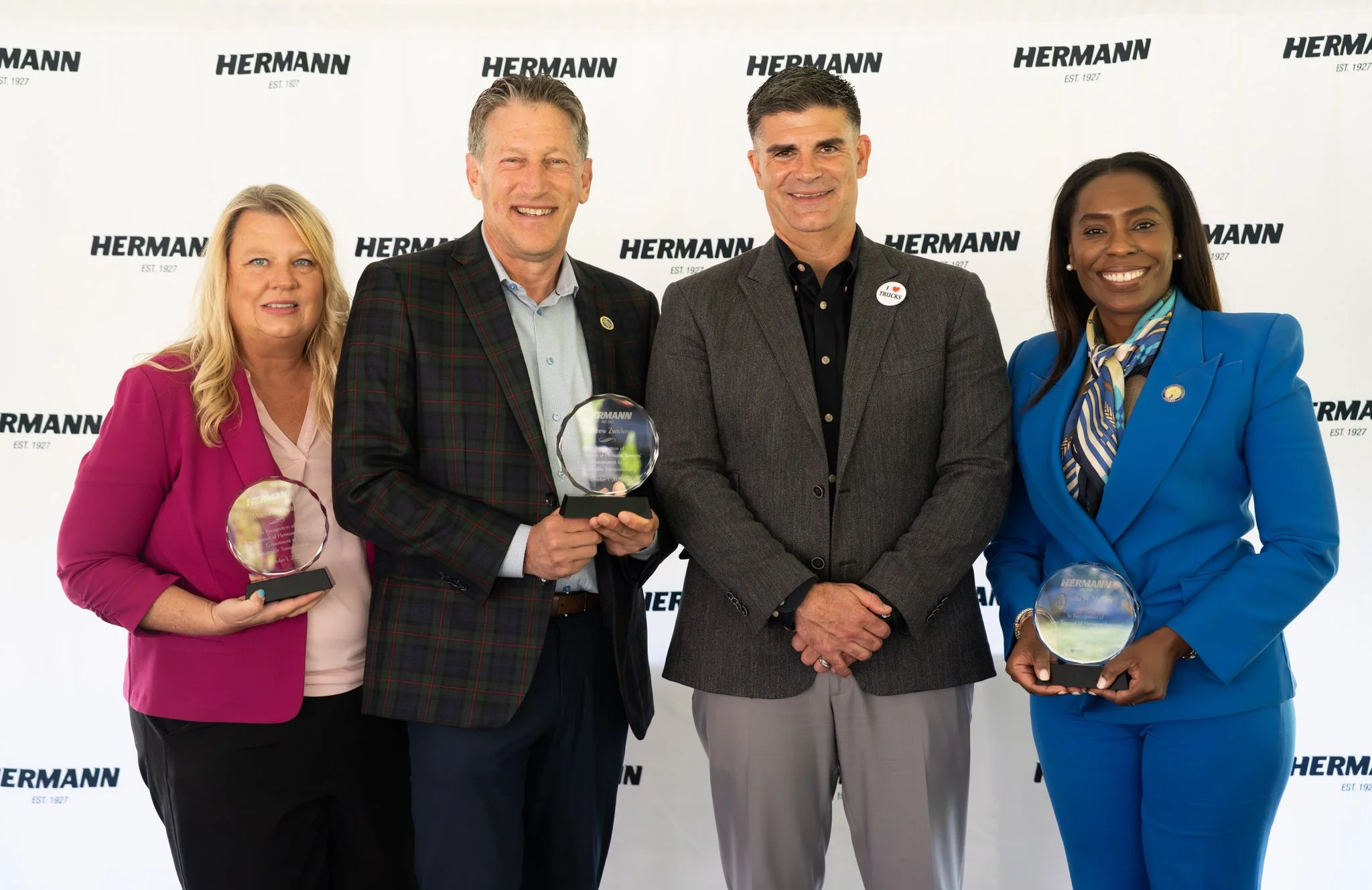 Four people standing in front of a Hermann branded backdrop, three of them holding glass awards, smiling for a photo. The group includes two women and two men, dressed in business attire.