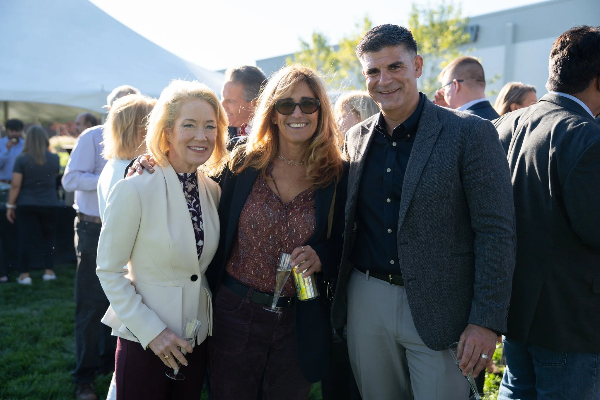 Three people smiling at an outdoor event holding glasses, with many other people in the background.