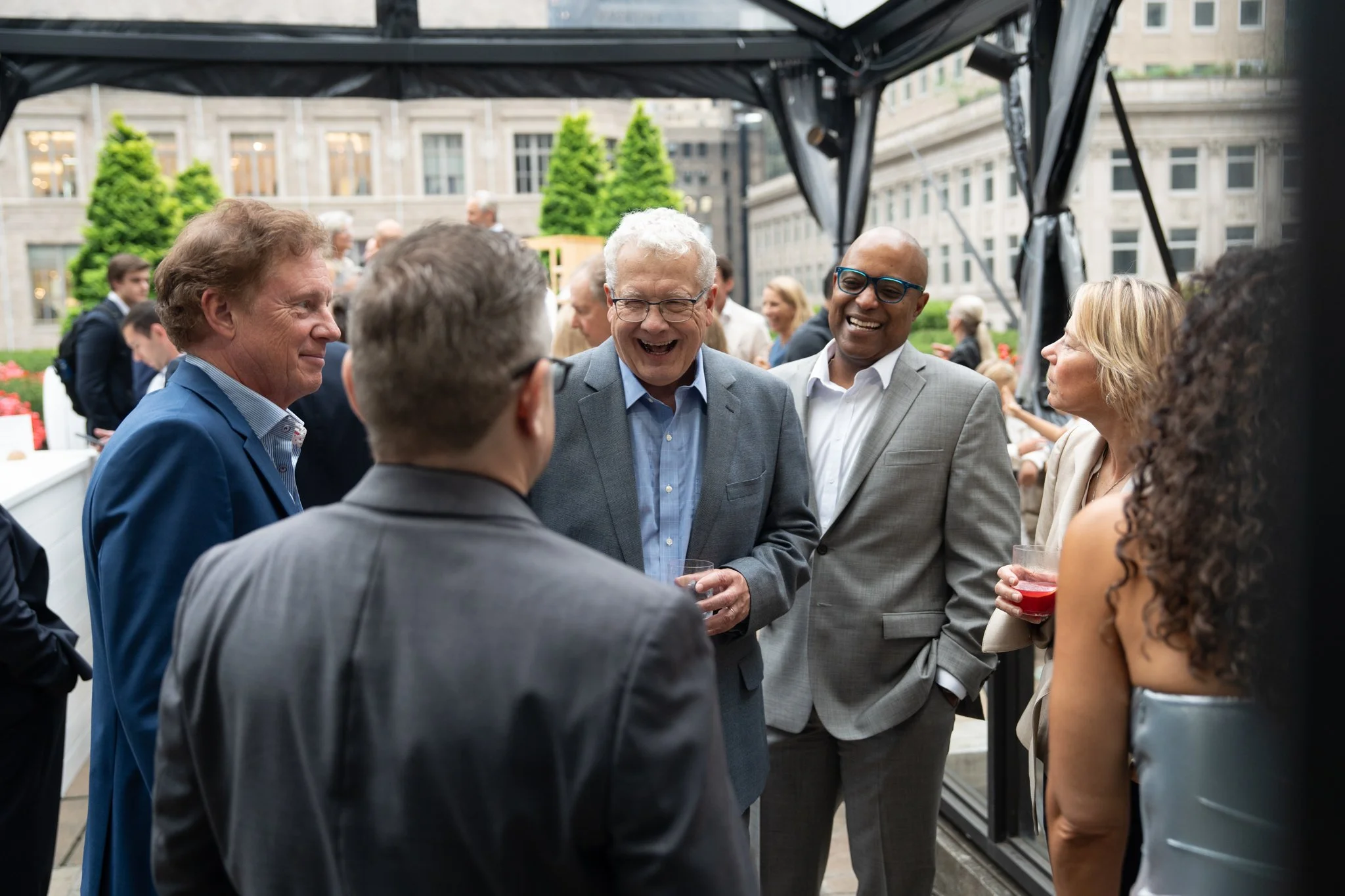 Group of professionally dressed people socializing and smiling at an outdoor event under a canopy, with a cityscape and greenery in the background.