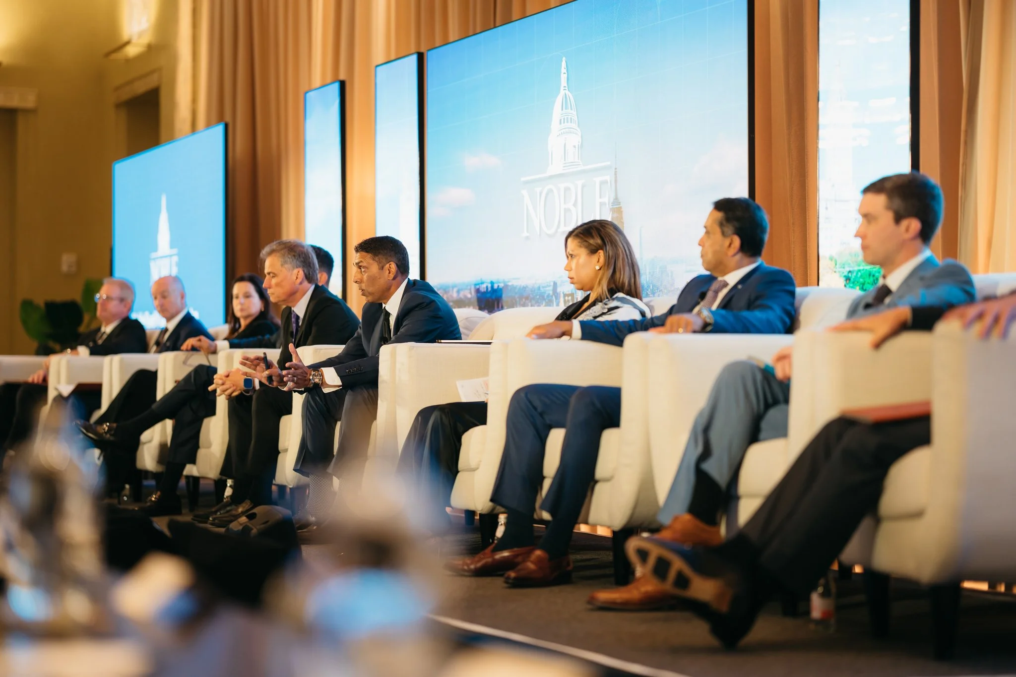 Group of people sitting on stage at a conference, with large screens in the background displaying the word 'Noble' and a building illustration.