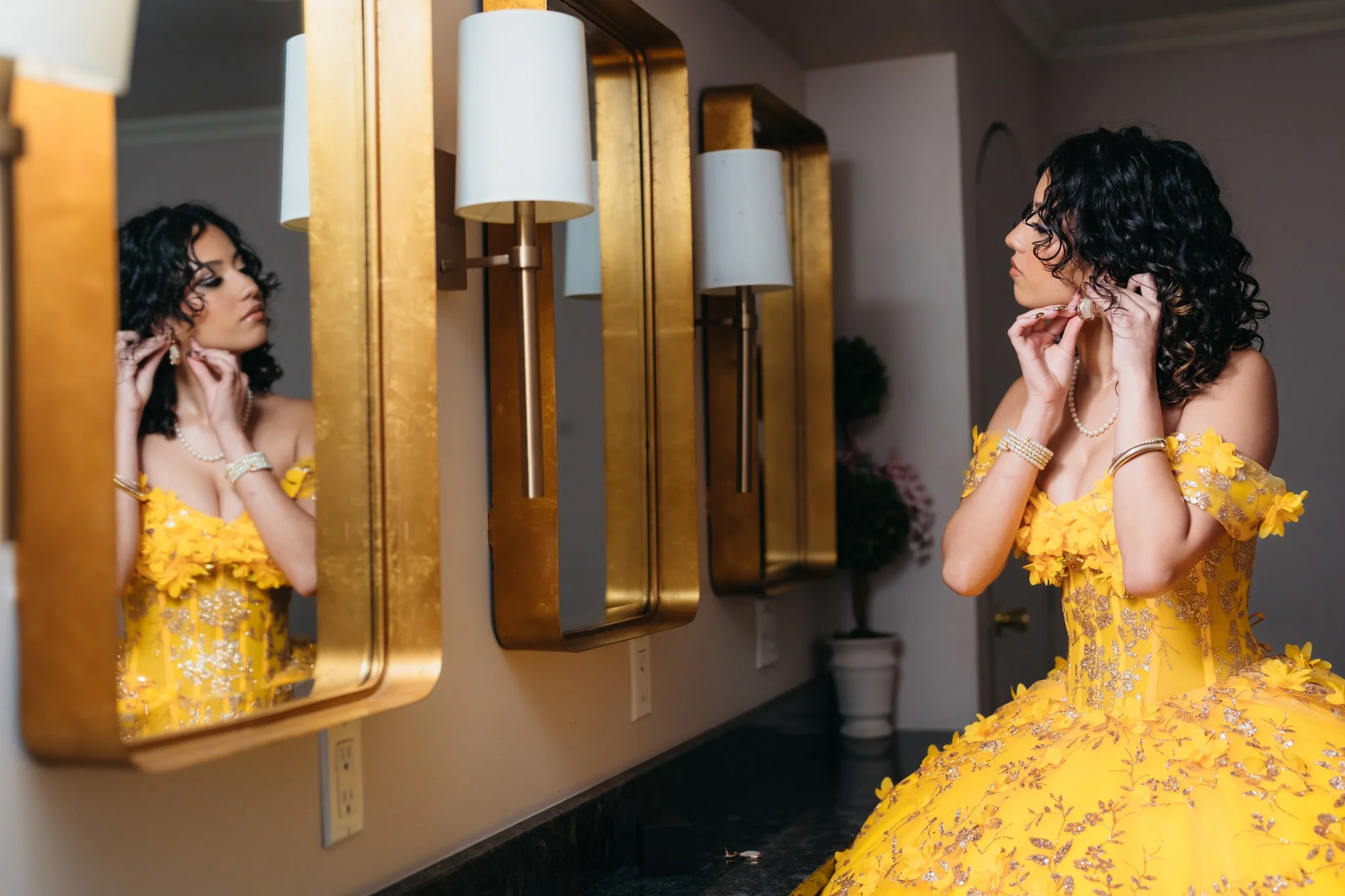 A woman in a bright yellow, floral, off-shoulder gown is looking at her reflection in a mirror as she adjusts her earrings. She has curly black hair and is wearing jewelry including a pearl necklace, bracelet, and earrings. The scene is set in an ele