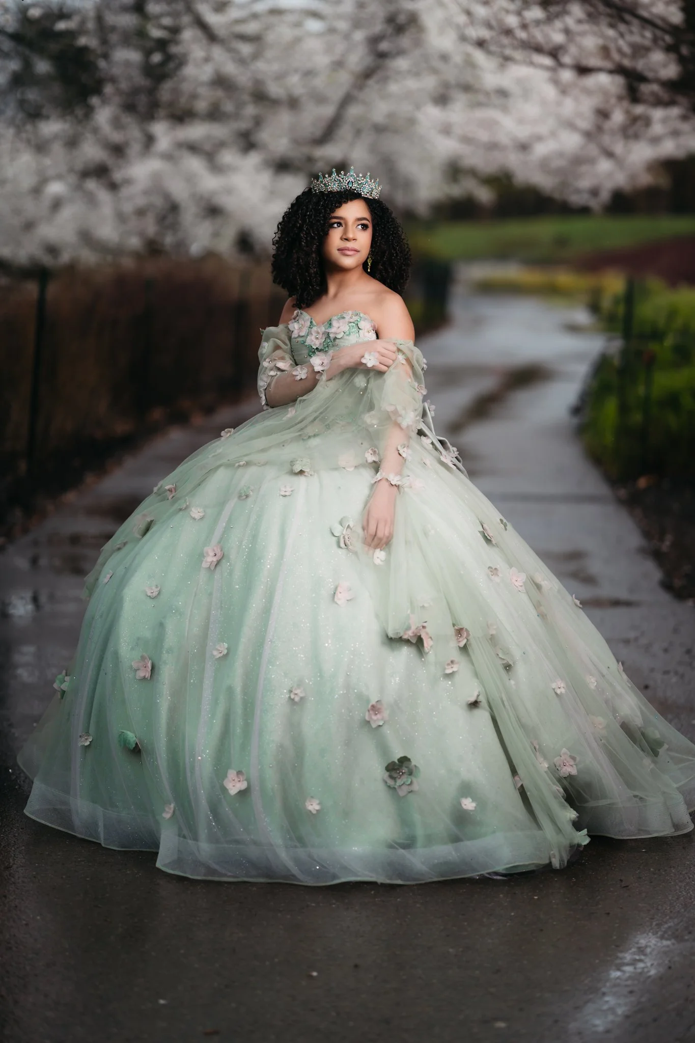 Young woman in an elaborate green gown decorated with pink flowers, wearing a crown, standing on a pathway with blossoming cherry trees in the background.