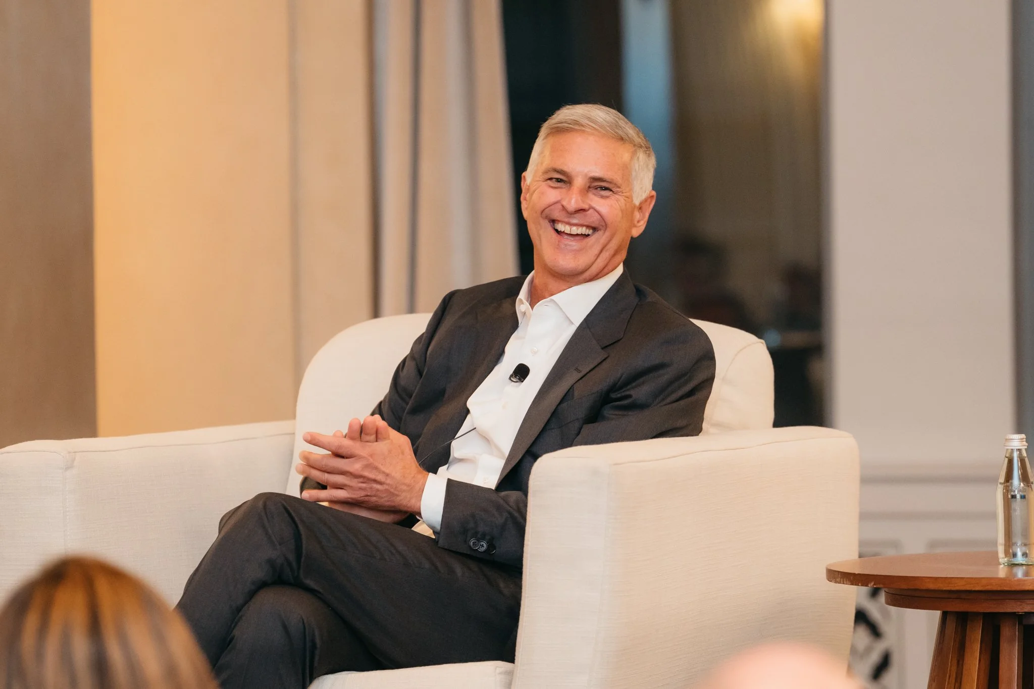 A smiling man in a dark suit and white shirt sitting on a white sofa during a speaking event, with a microphone clipped to his shirt.