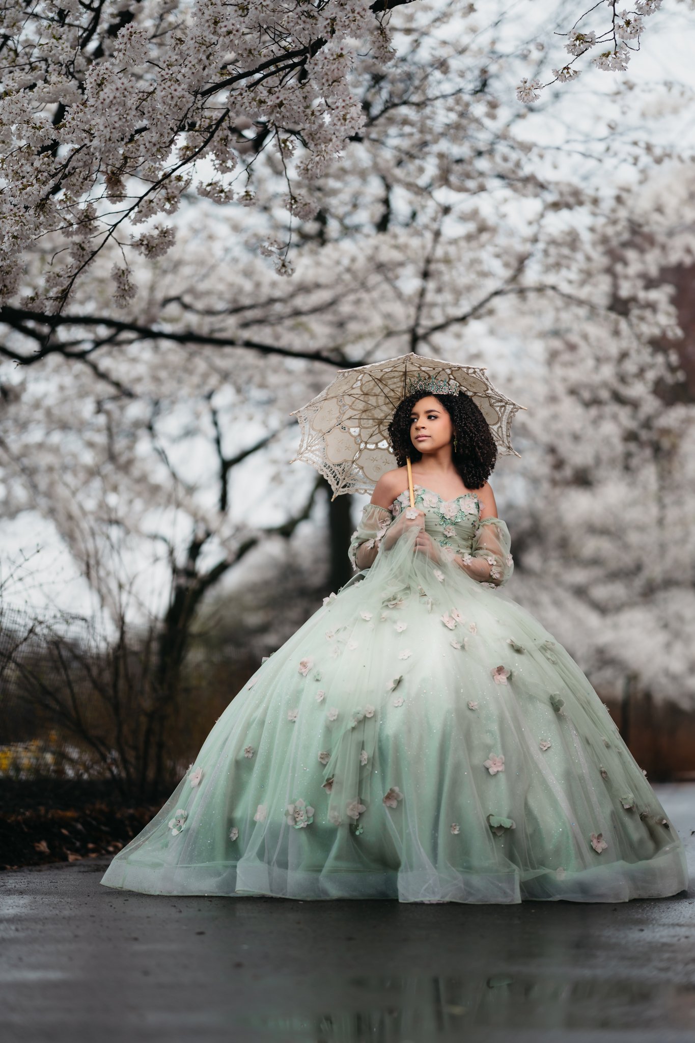 A woman in a pale green, floral ball gown standing outdoors, holding a lace parasol, surrounded by blooming cherry blossom trees.