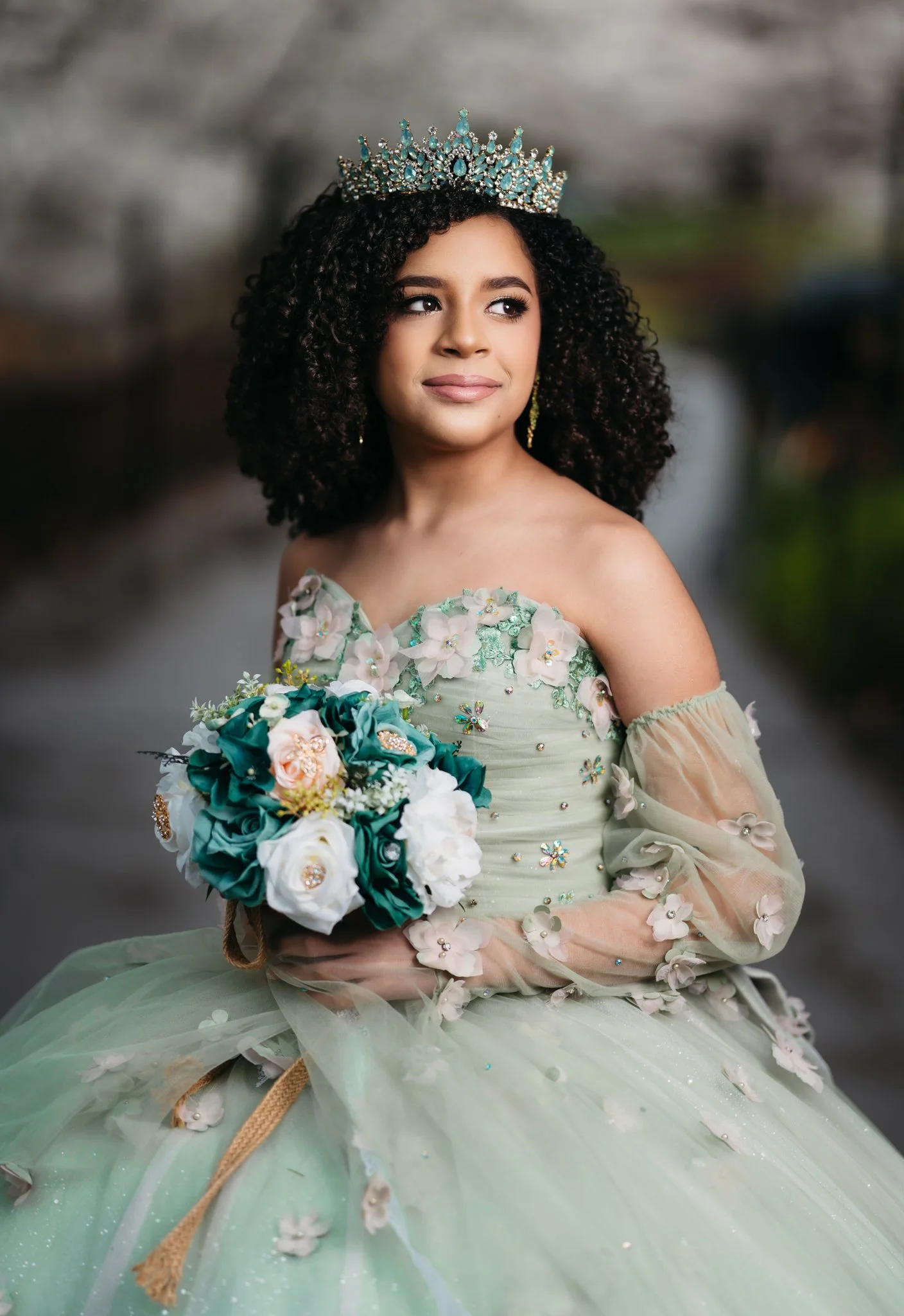 A young woman in a pastel green dress decorated with 3D floral appliqués and embellishments, wearing a large tiara, holding a bouquet of roses and greenery, with a blurred outdoor background.