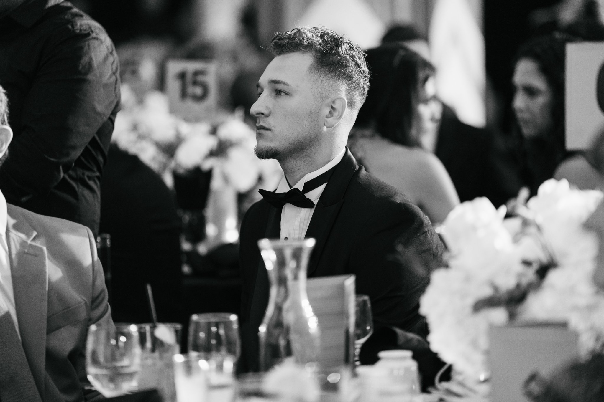 A young man in a tuxedo with a bow tie, seated at a formal event table, looking to the side with a serious expression. The table is decorated with flowers, glasses, and a vase, and there are other seated guests in the background.