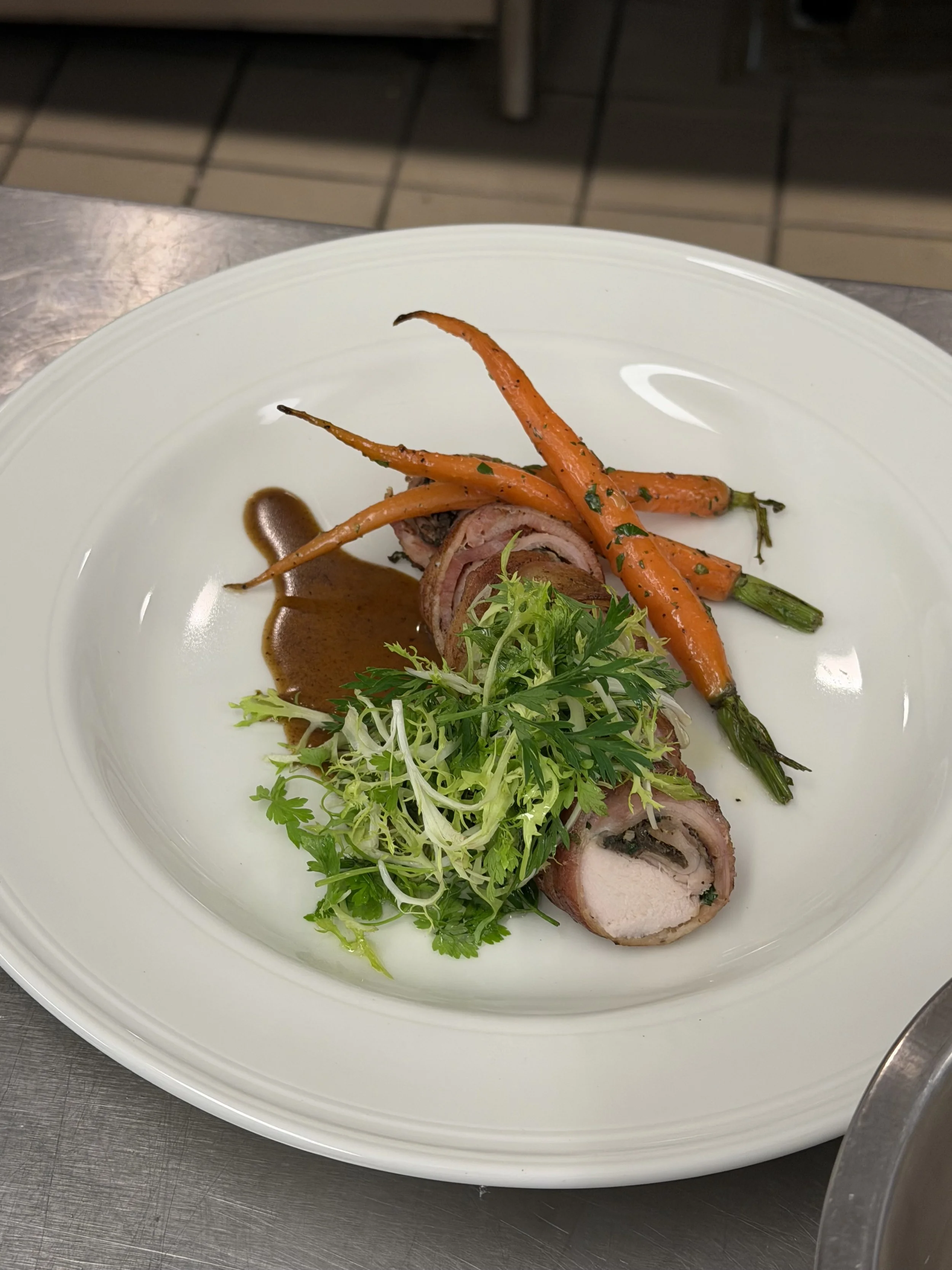 A plated dish featuring sliced stuffed meat, roasted carrots with herbs, and a side of mixed greens, garnished with a brown sauce on a white plate.
