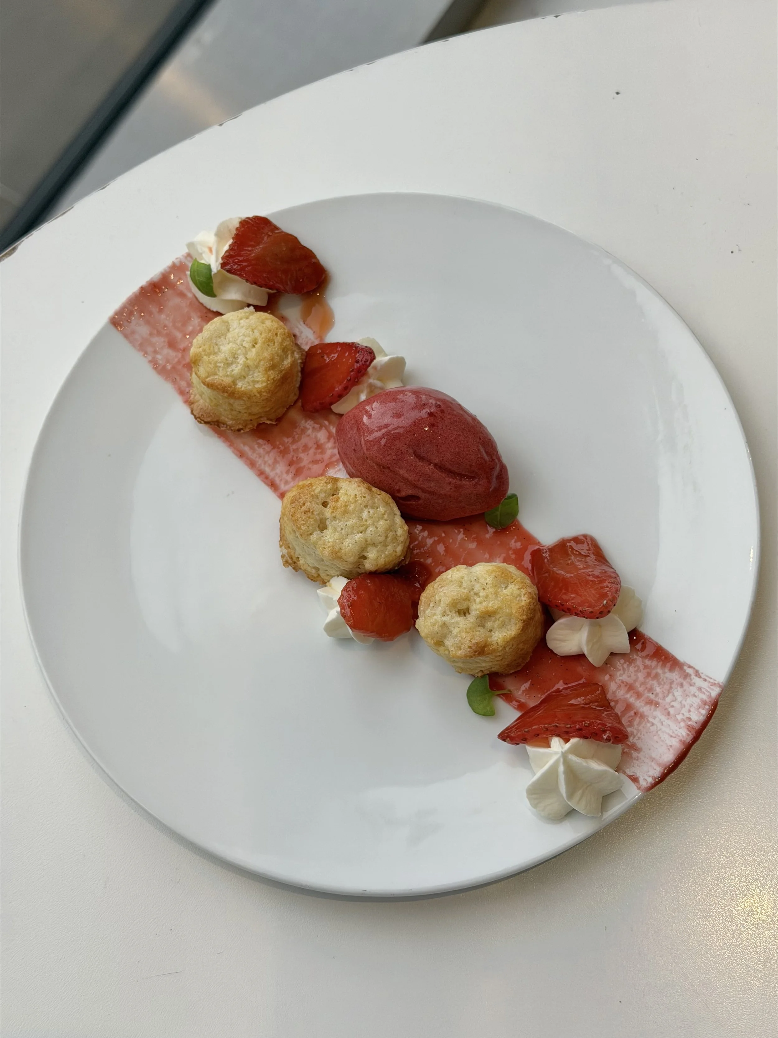 A white plate of dessert featuring strawberries, cream, small pastries, green leaves, and a red sauce on a white surface.