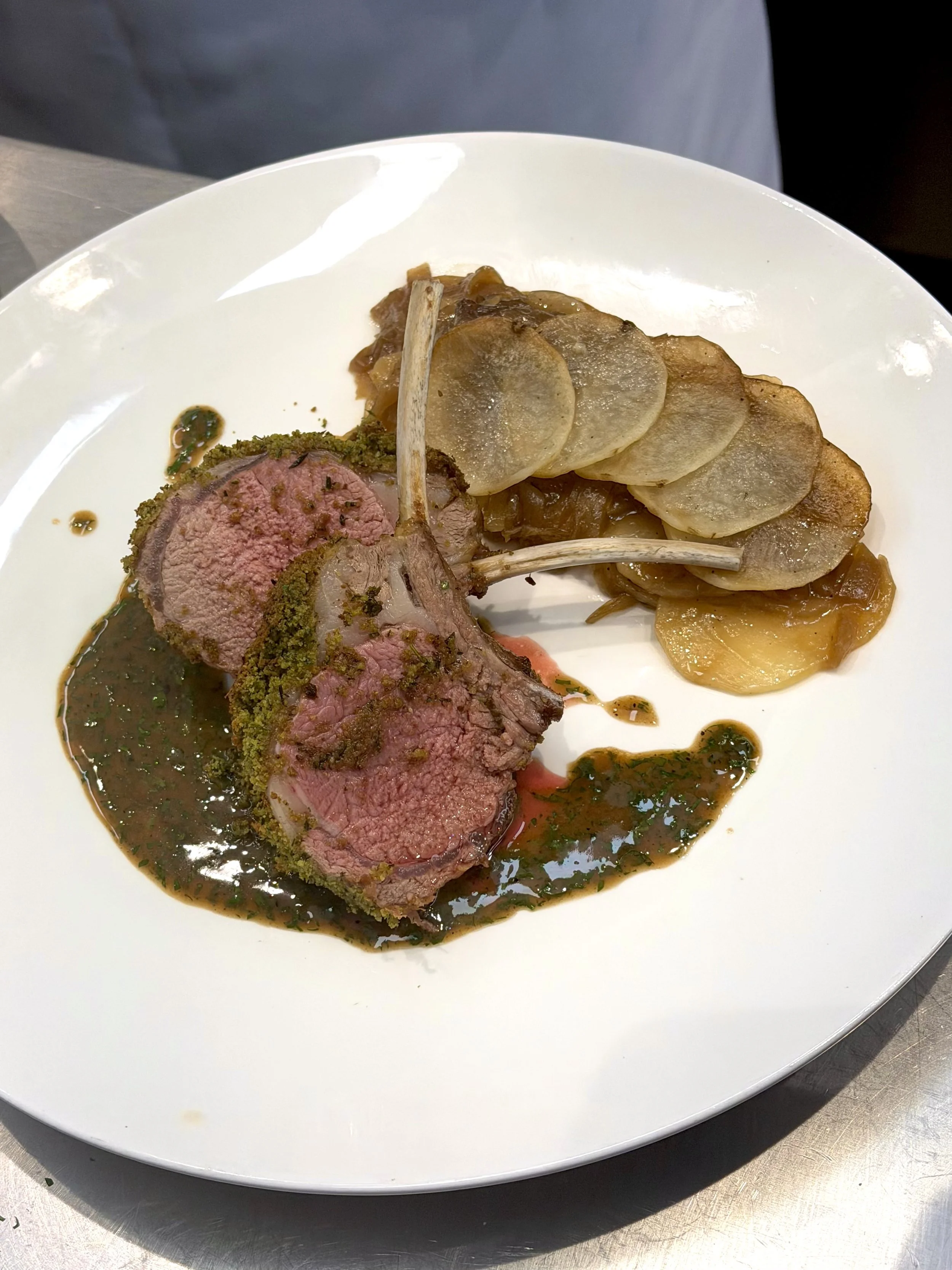 A plate with sliced roasted lamb, potato chips, and a serving of cooked vegetables, garnished with a green herb sauce.