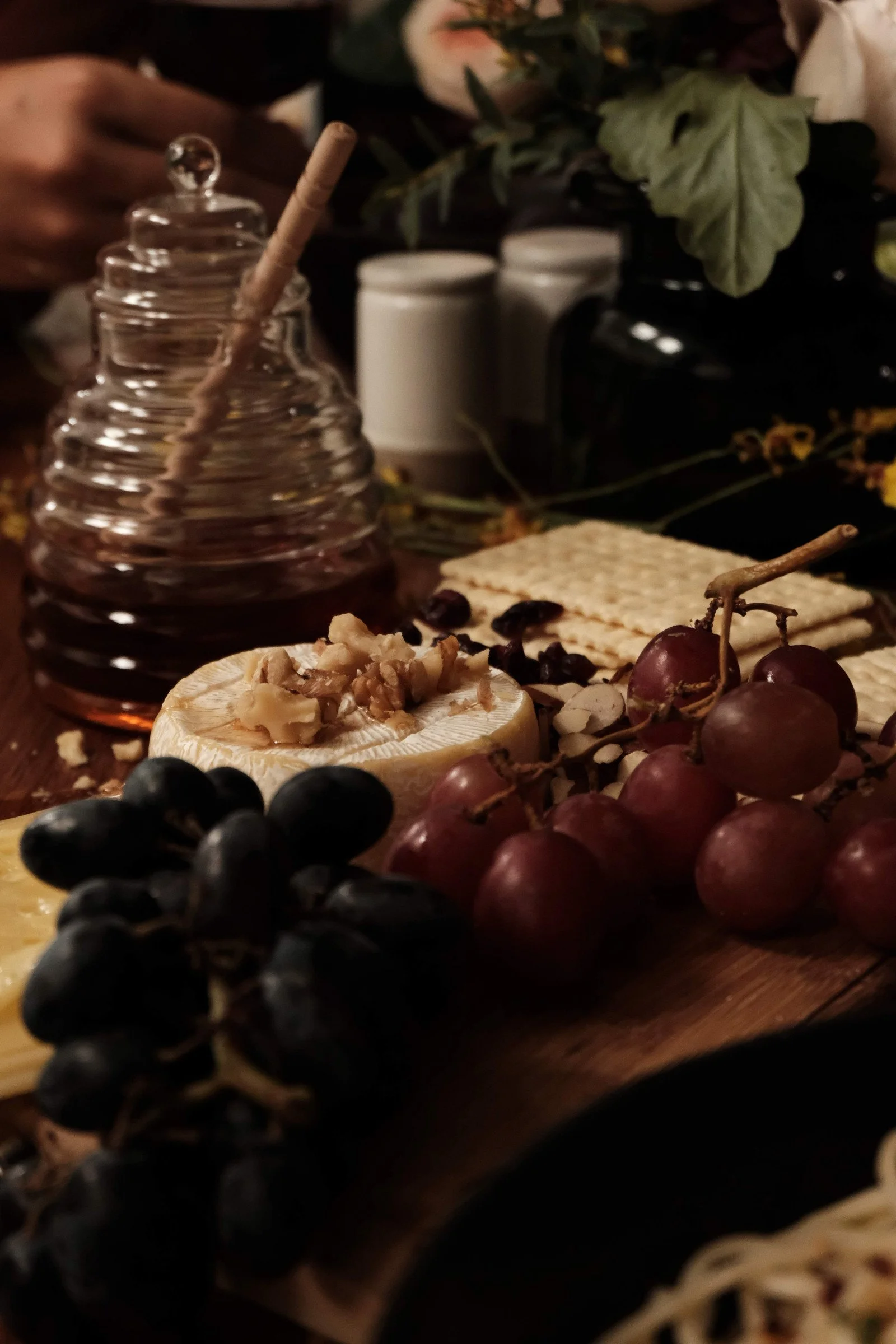A variety of cheeses, fresh grapes, crackers, nuts, honey jar, and a bunch of grapes on a wooden board.