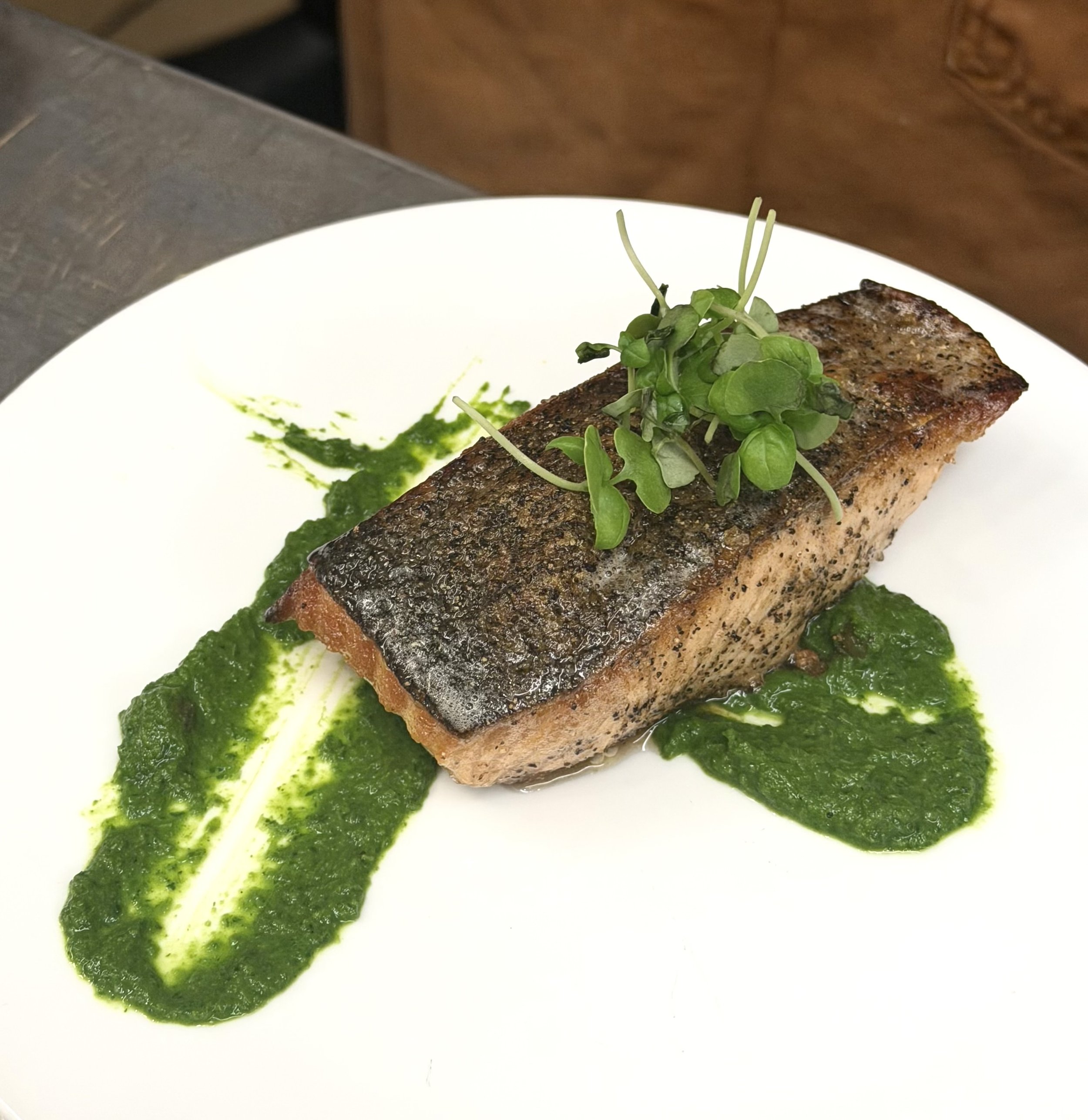 A piece of grilled salmon topped with microgreens, served on a white plate with green sauce.