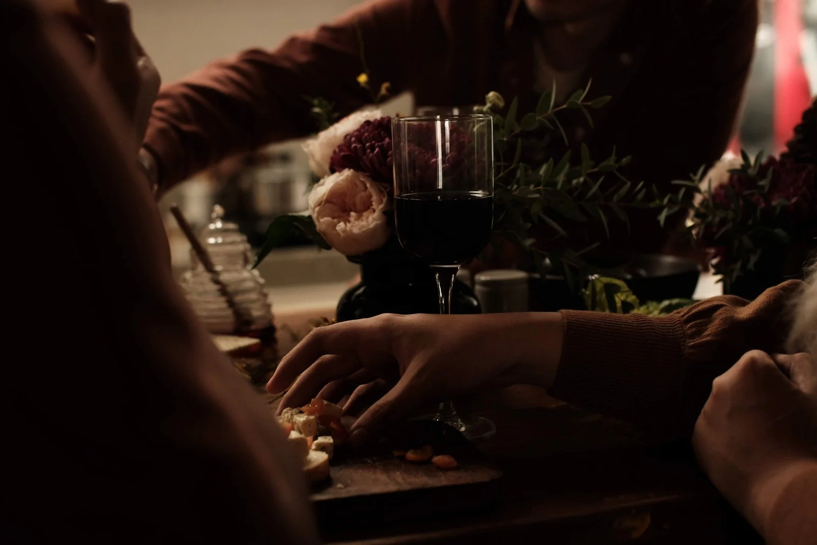 People sharing a meal and drink in a dimly lit setting, with a glass of red wine and a floral centerpiece visible.