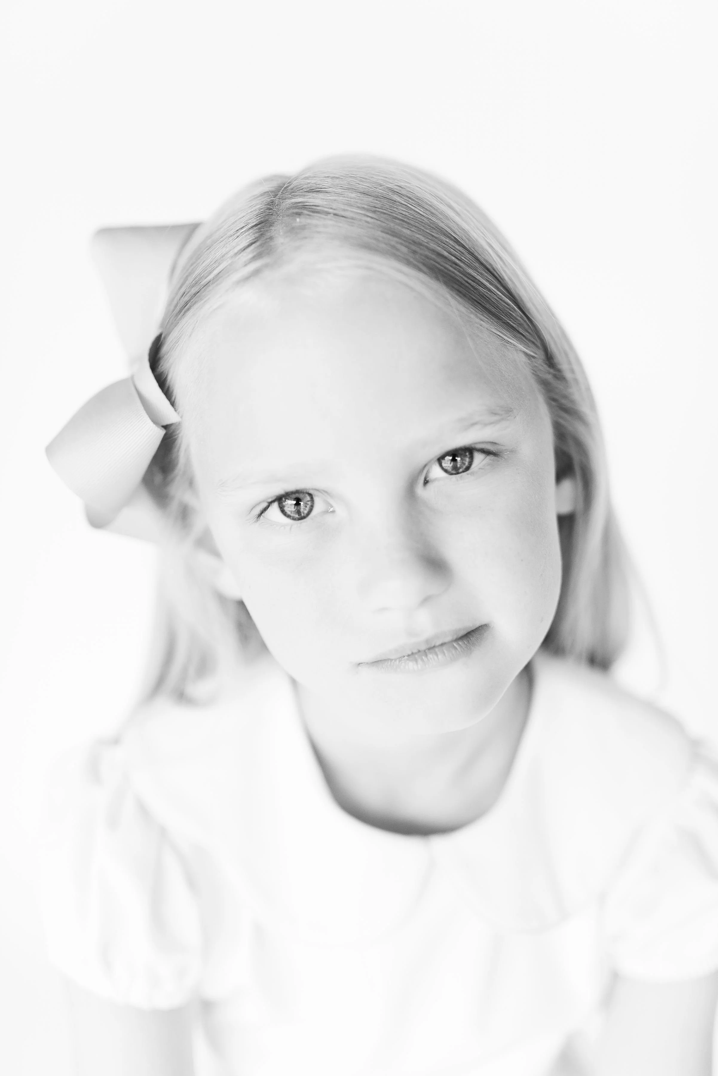 Black and white close-up photo of a child with light-colored hair, gazing directly into the camera, wearing a white shirt and a bow in hair.