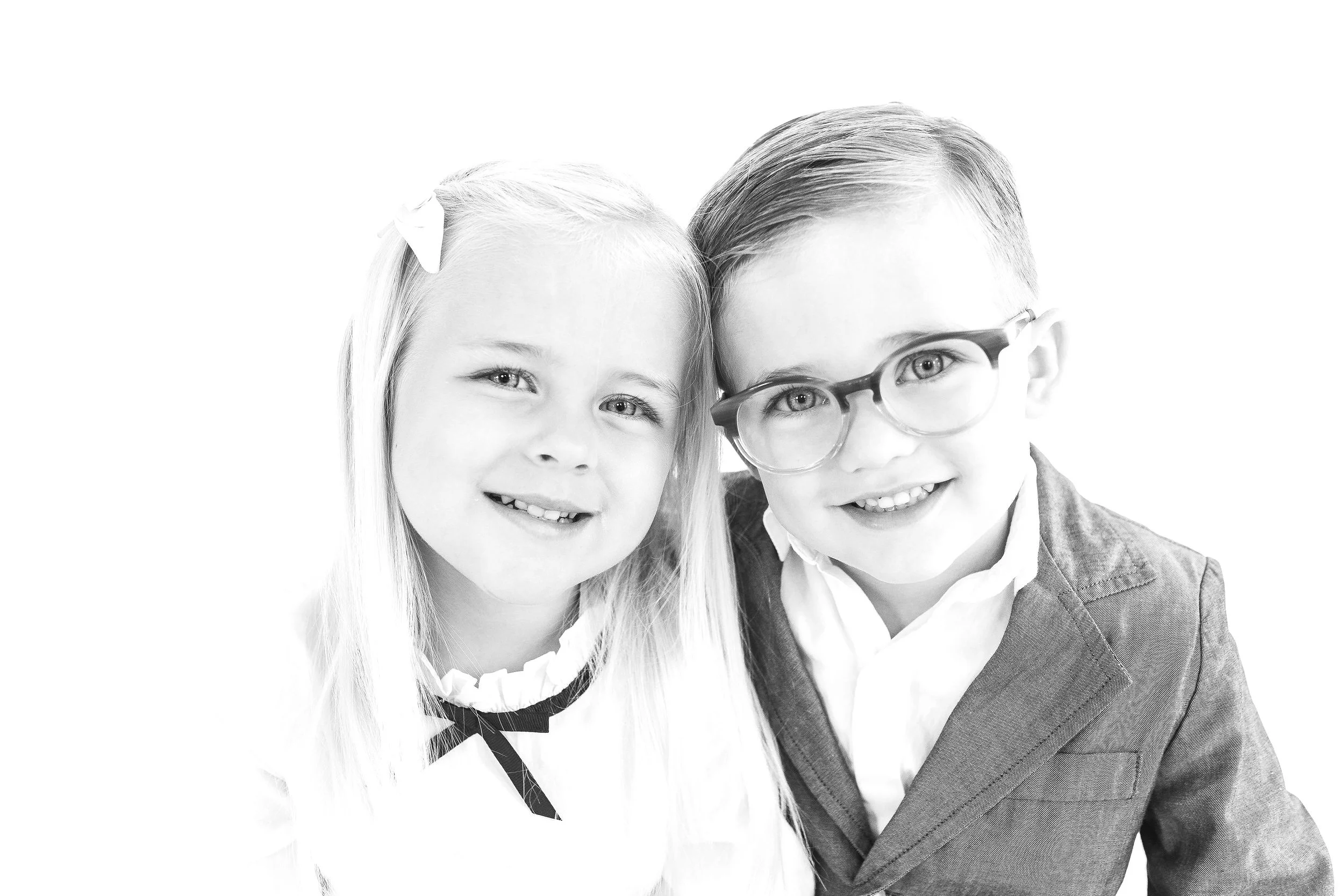 A black and white photo of a young girl with long hair and a bow, and a young boy with glasses, both smiling and close together.