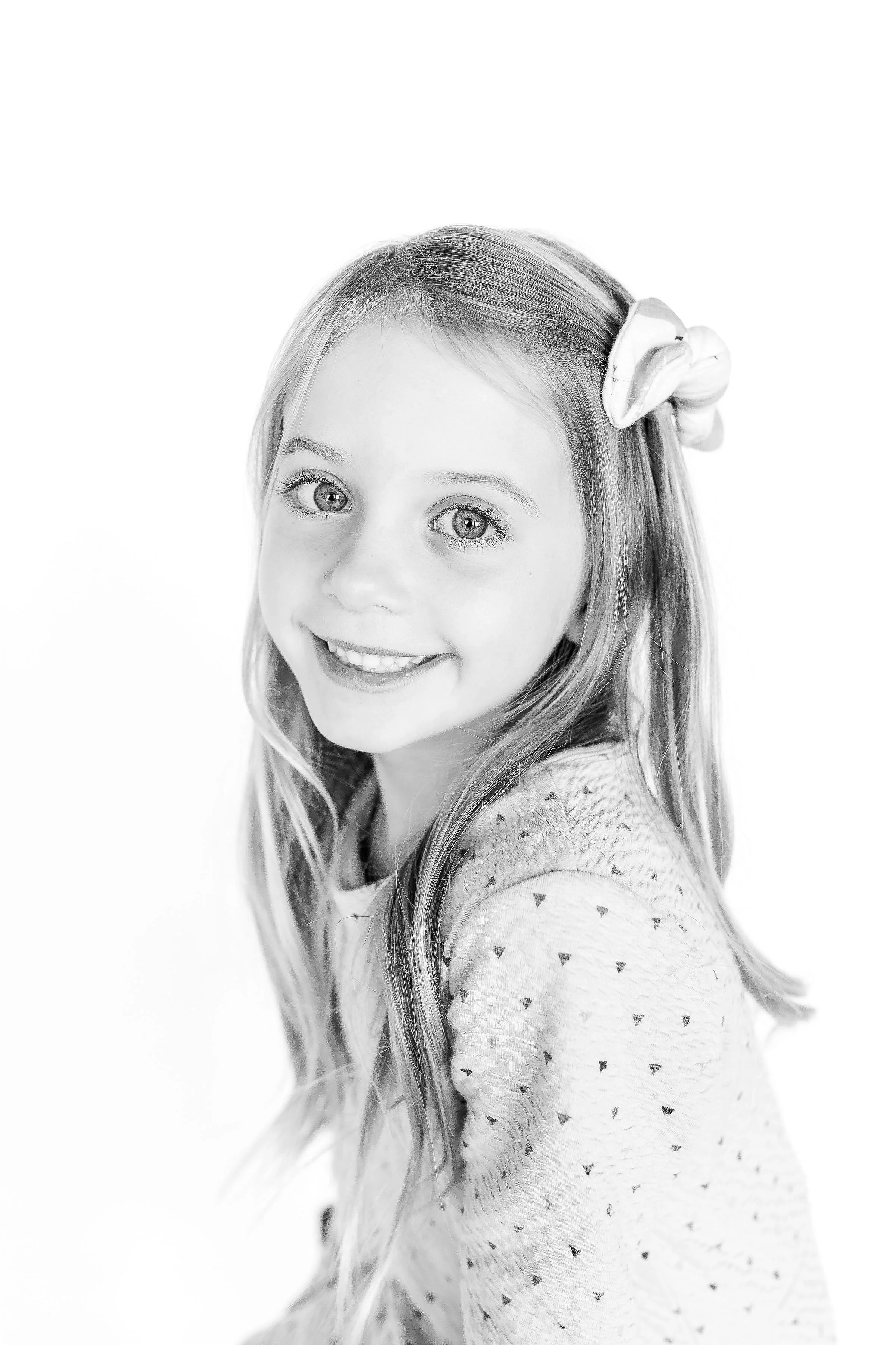 Black and white portrait of a young girl with long hair, bright eyes, and a big smile, wearing a sweater with small heart patterns, and a large flower hair accessory.