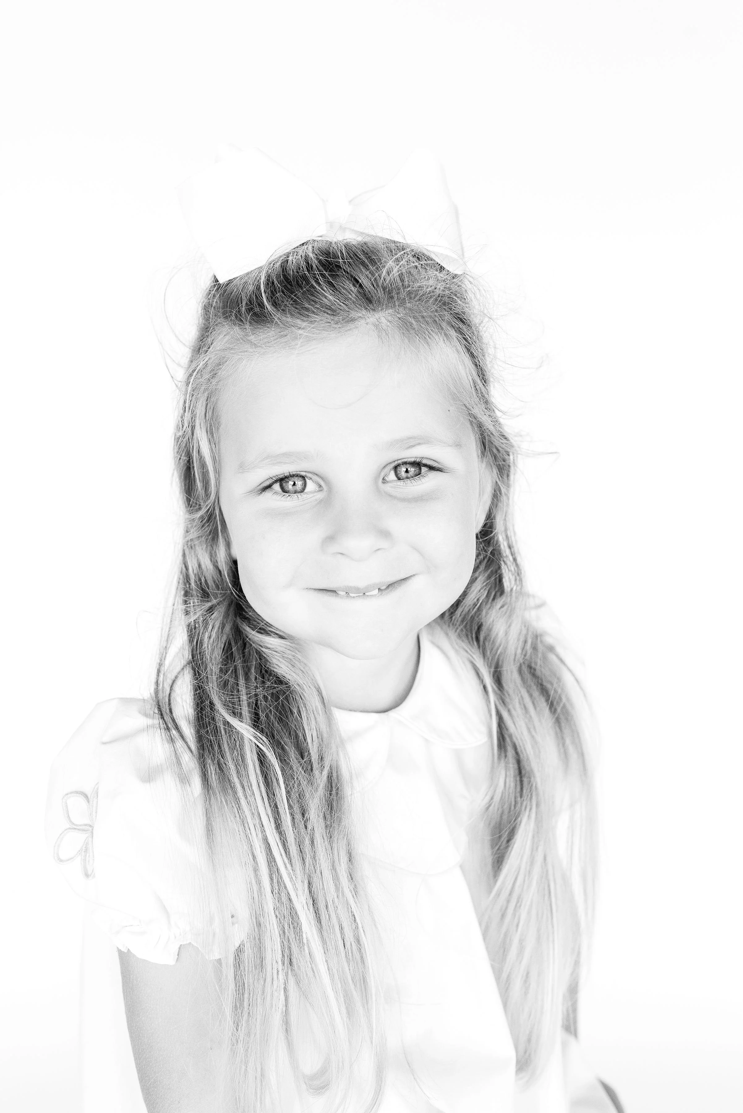 A young girl with long hair and a large bow in her hair, smiling at the camera, wearing a light-colored top with short puffed sleeves, in a high-key black and white photograph.