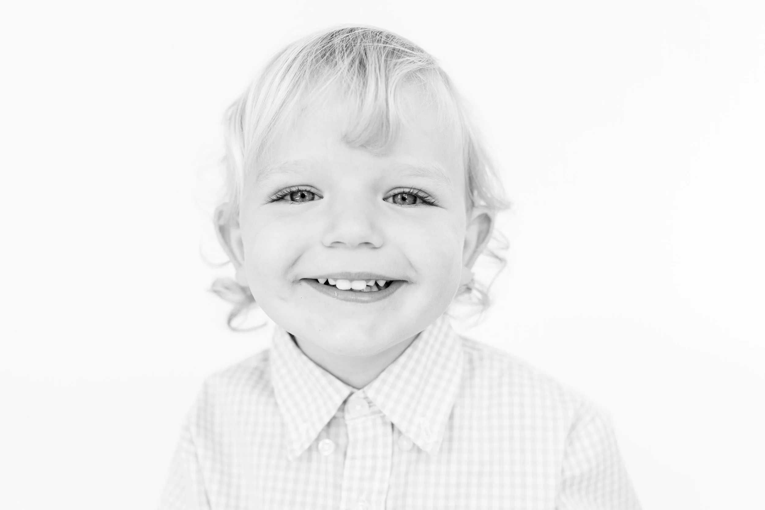 Black and white portrait of a smiling young child with light-colored hair, wearing a checked shirt.