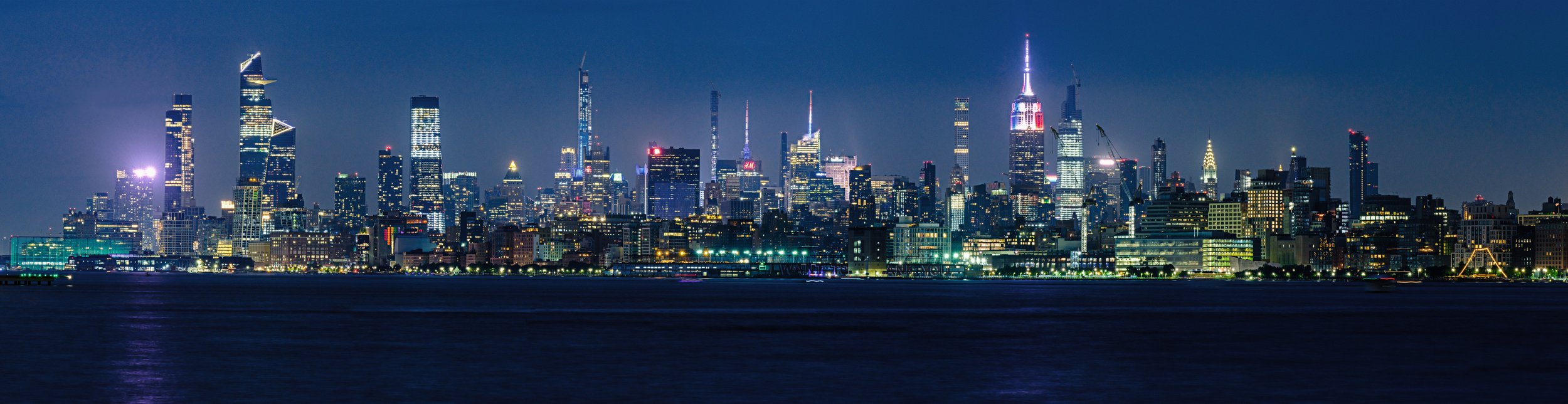 Panoramic view of the Midtown East skyline of NYC at night, including Hudson Yards, the Empire State Building, and the Chrysler Building