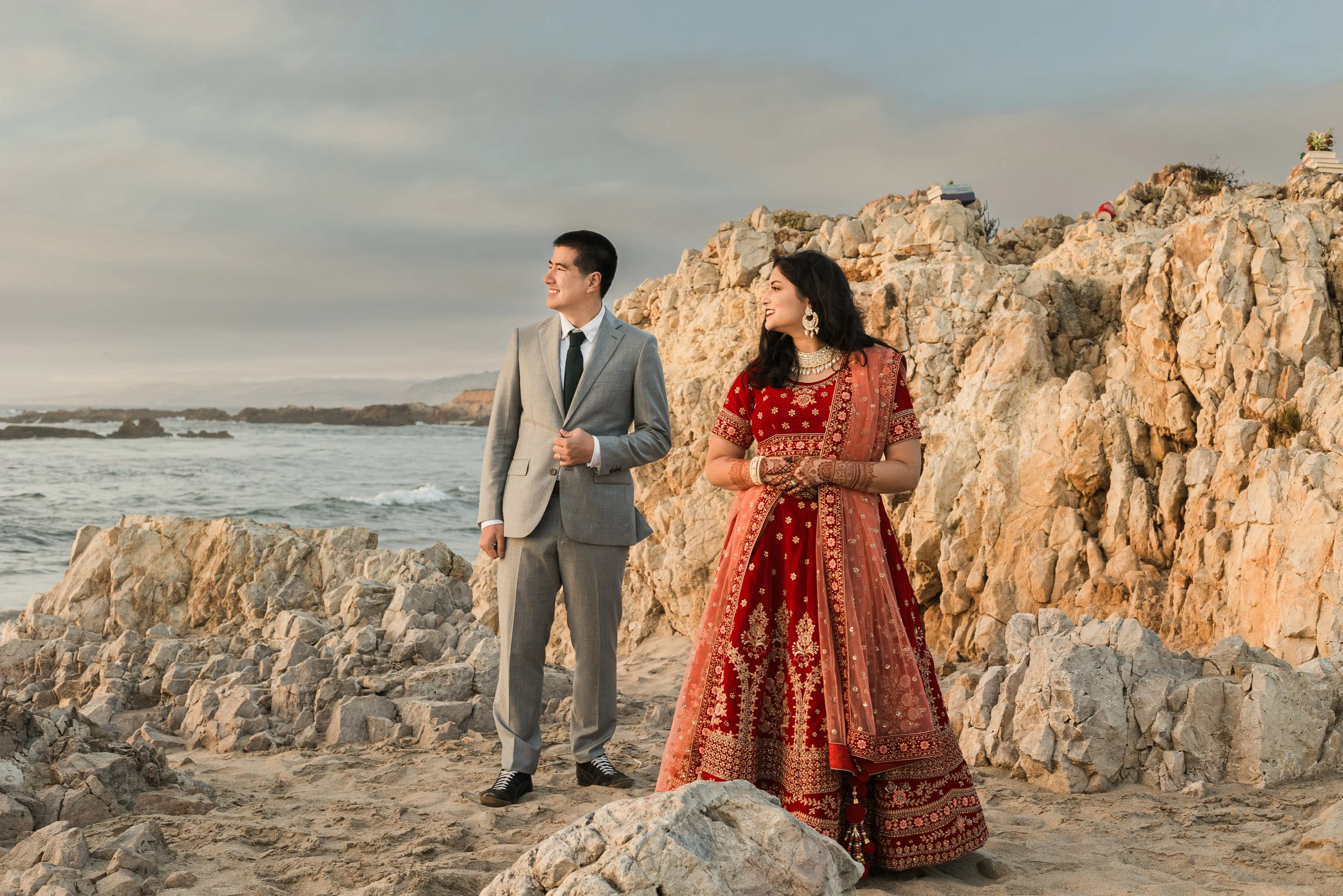 Groom in suit and bride in traditional Indian dress stand on a rocky beach in Pacifica.