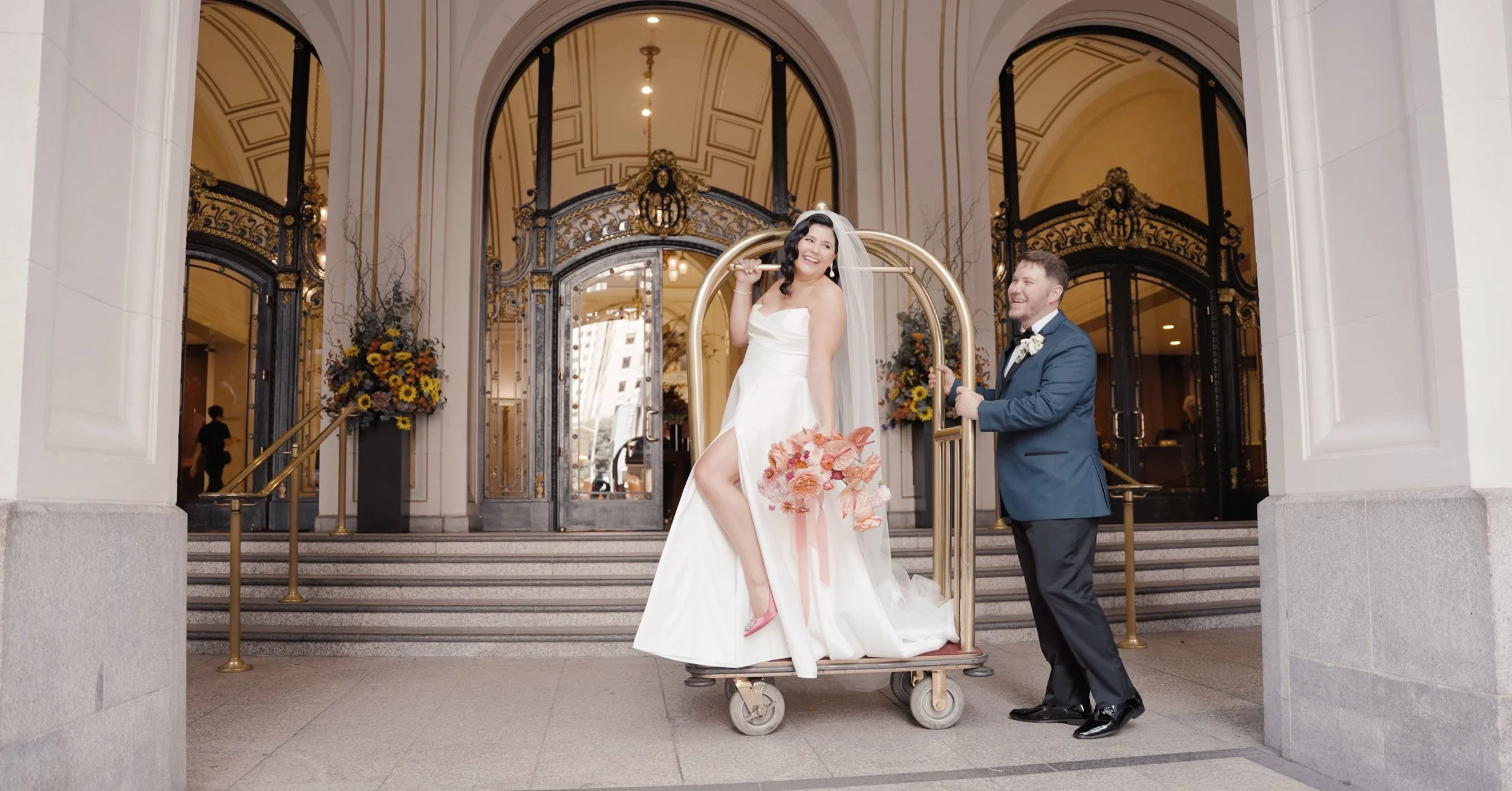 Bride stands on luggage cart while the groom pushes it at the Palace Hotel San Francisco.