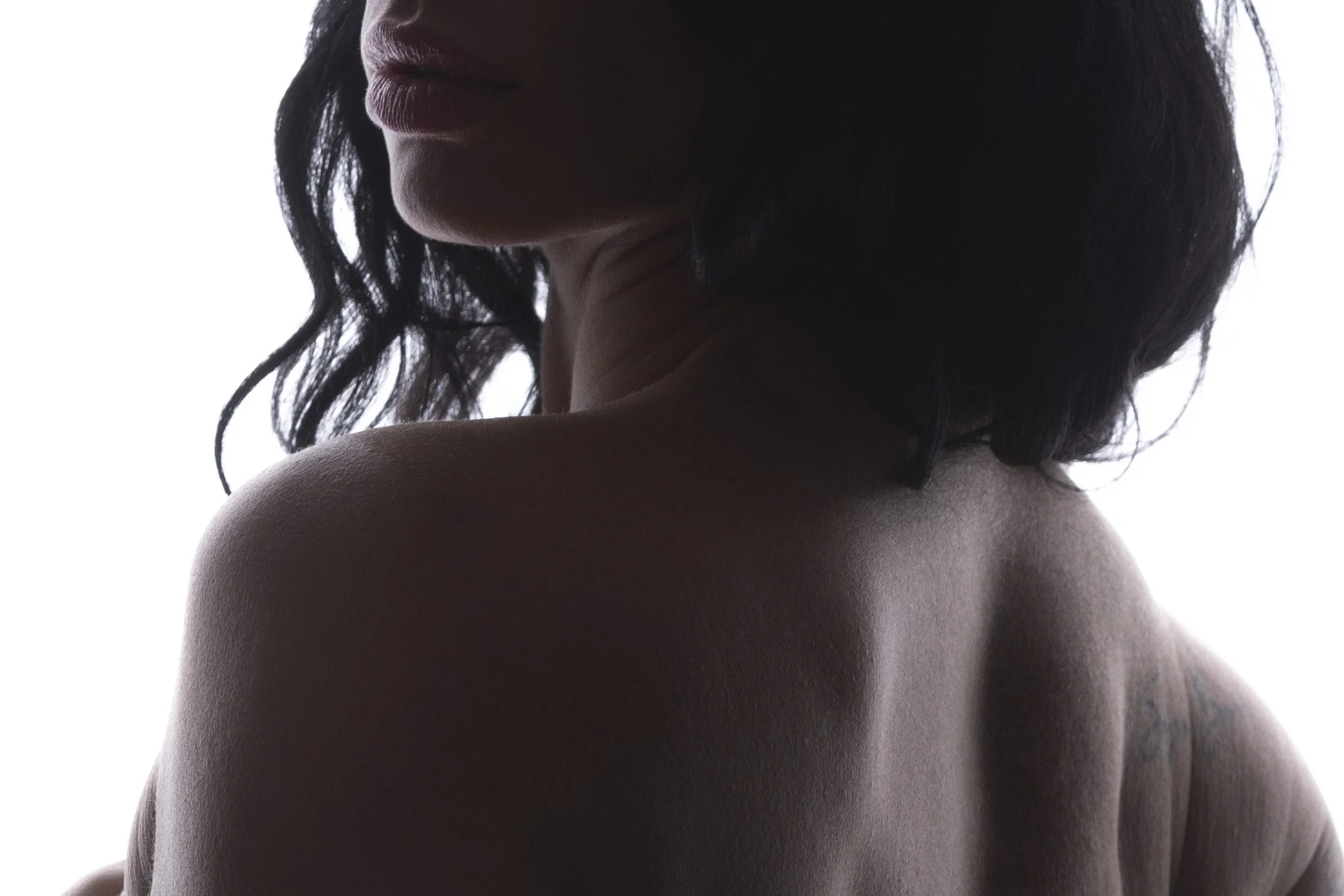 Close-up silhouette of a woman with dark, wavy hair, facing left, showing her shoulder and part of her face, against a white background.