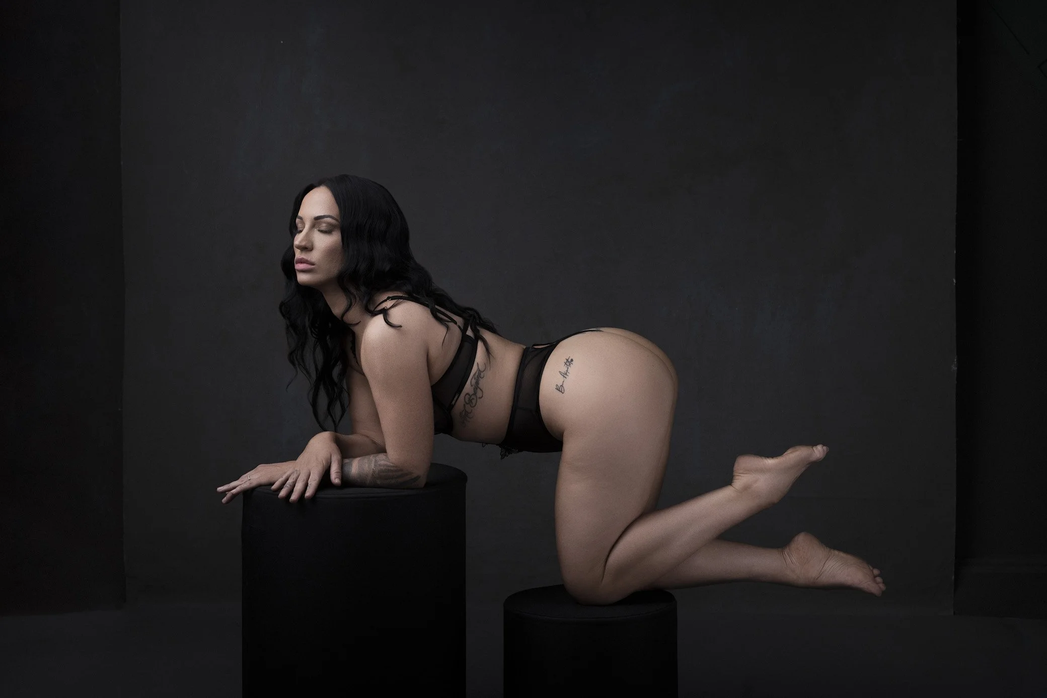 A woman with tattoos, dark hair, and minimal clothing poses on black cylindrical platforms against a dark background, with her eyes closed and her head turned to the side.