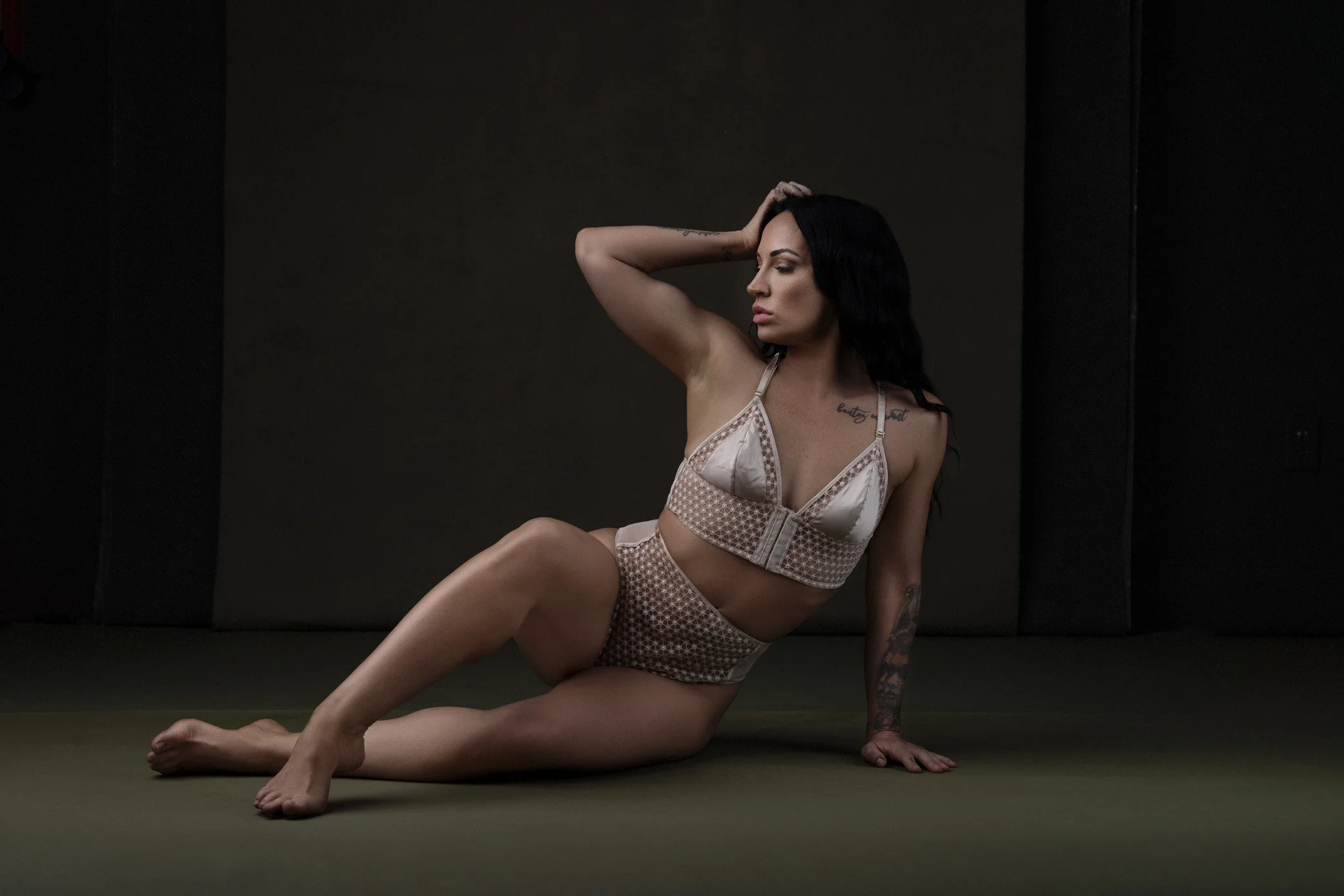 A woman with long dark hair and tattoos on her arm and chest, wearing beige patterned lingerie, sitting on the floor with one leg bent and the other extended, posing with one hand on her head and the other on the floor, against a dark background.