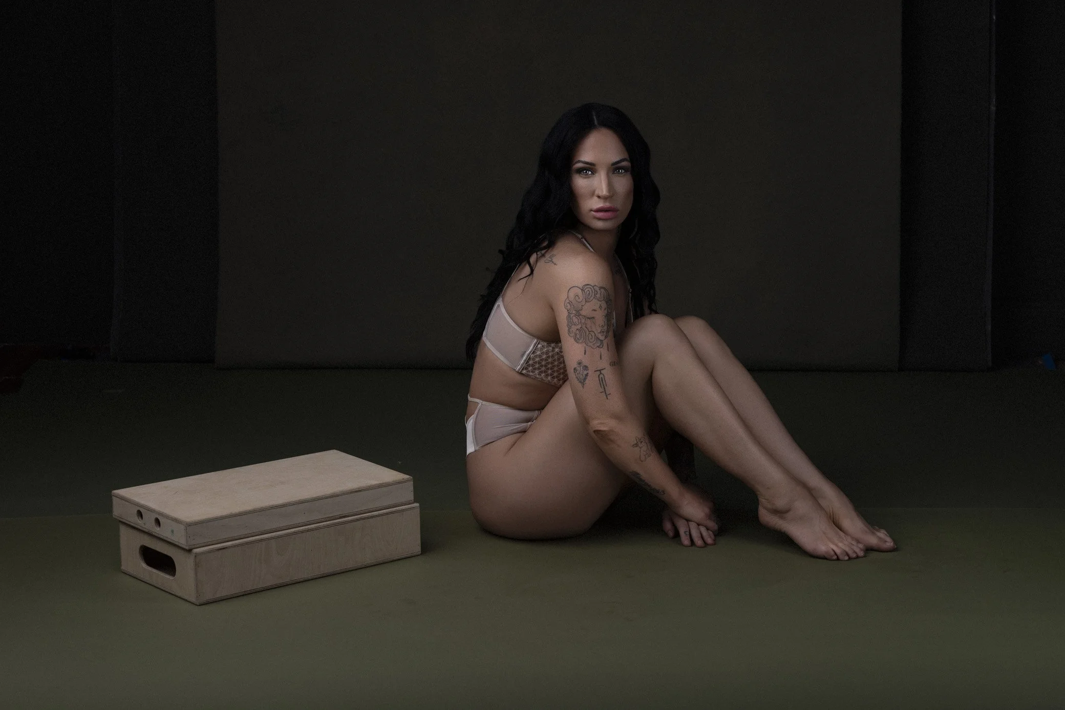 A woman with black hair and tattoos sitting on the floor in lingerie, with a wooden box nearby, in a dark room.