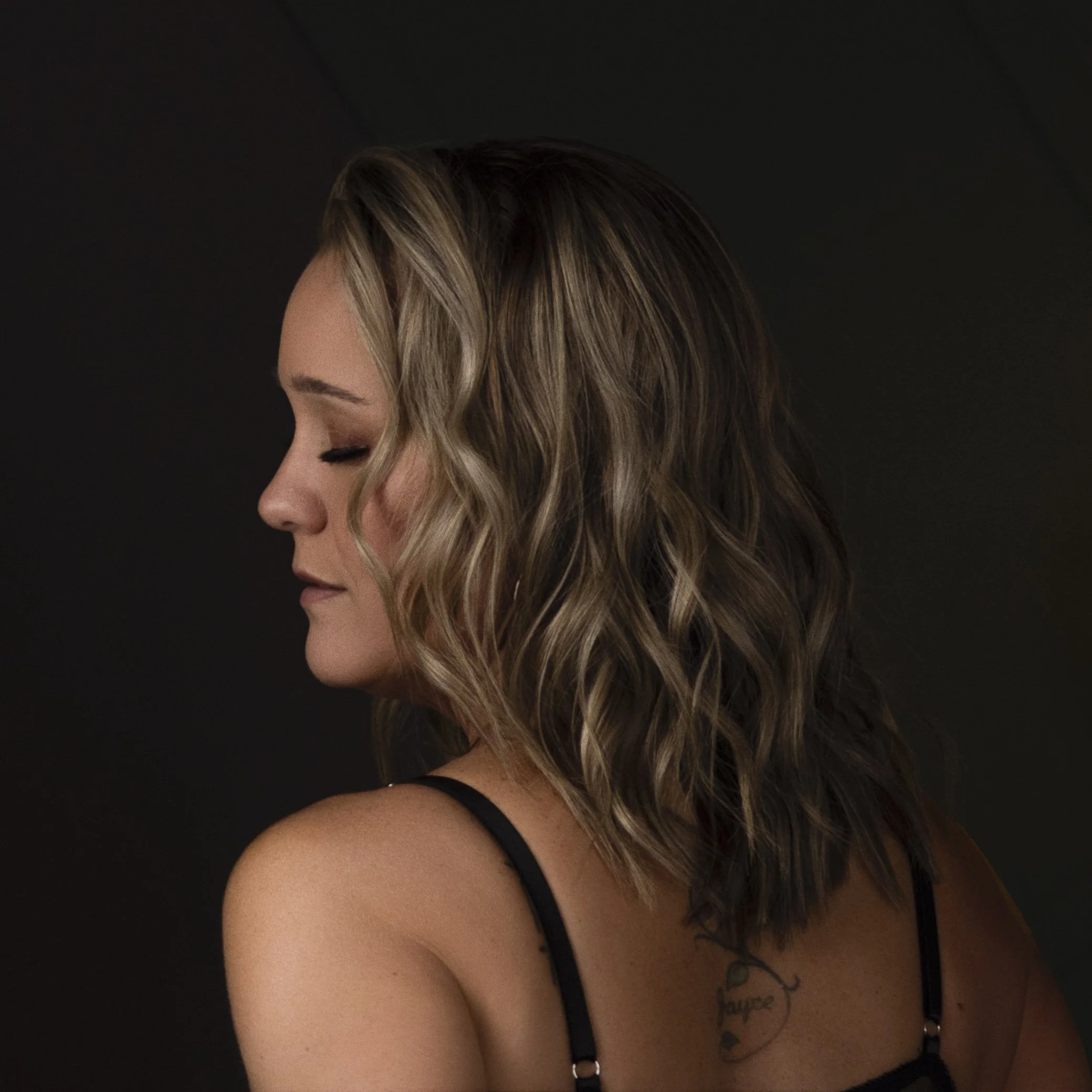 Profile of a woman with wavy blonde hair, eyes closed, wearing a black strap top, and a tattoo on her shoulder.