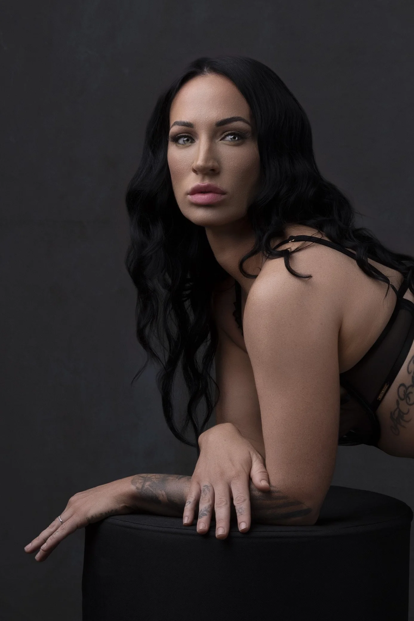 A woman with long black hair and tattooed arms looking at the camera against a dark background.