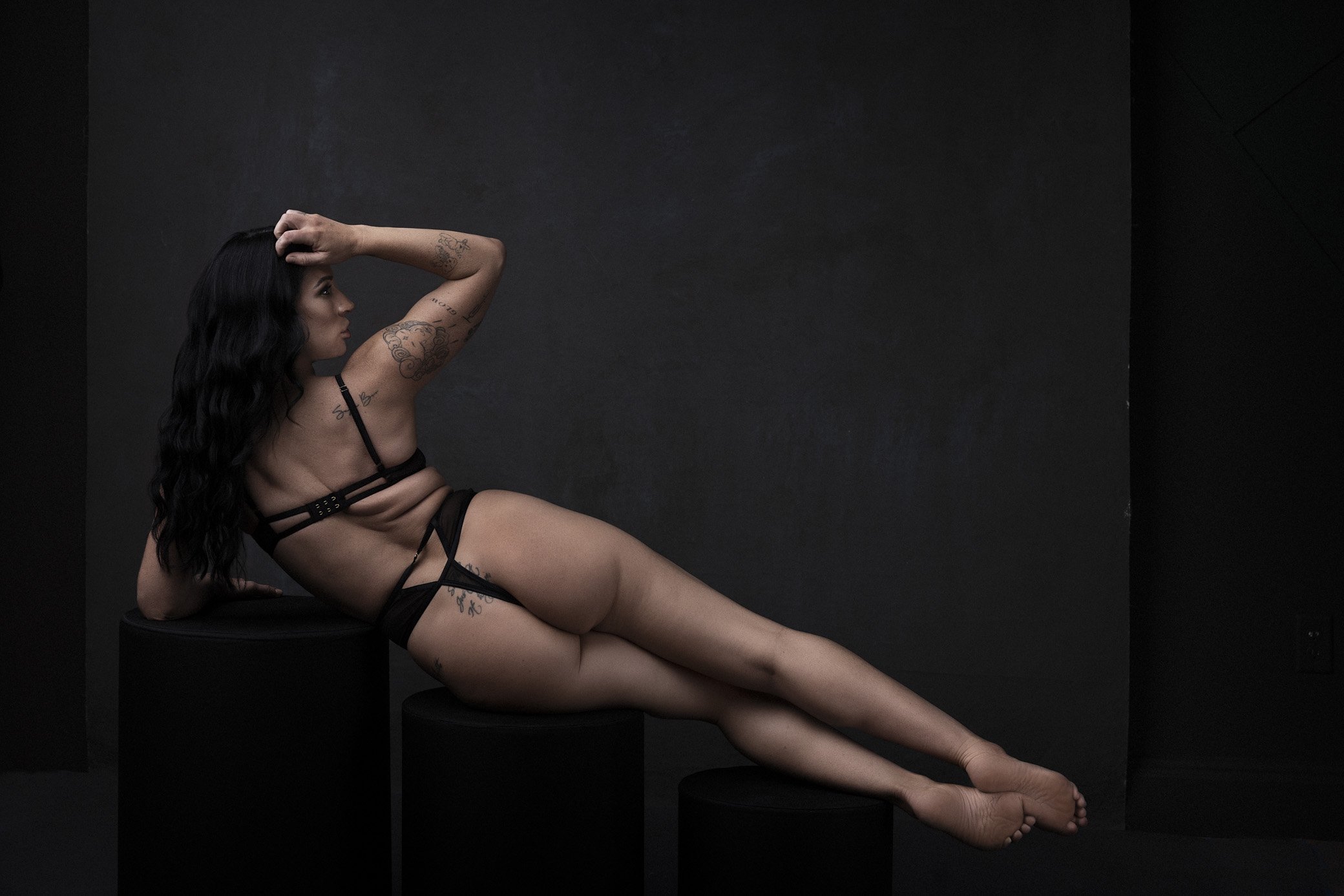 A woman with tattoos reclining on black cylinders set against a dark background, wearing black lingerie.