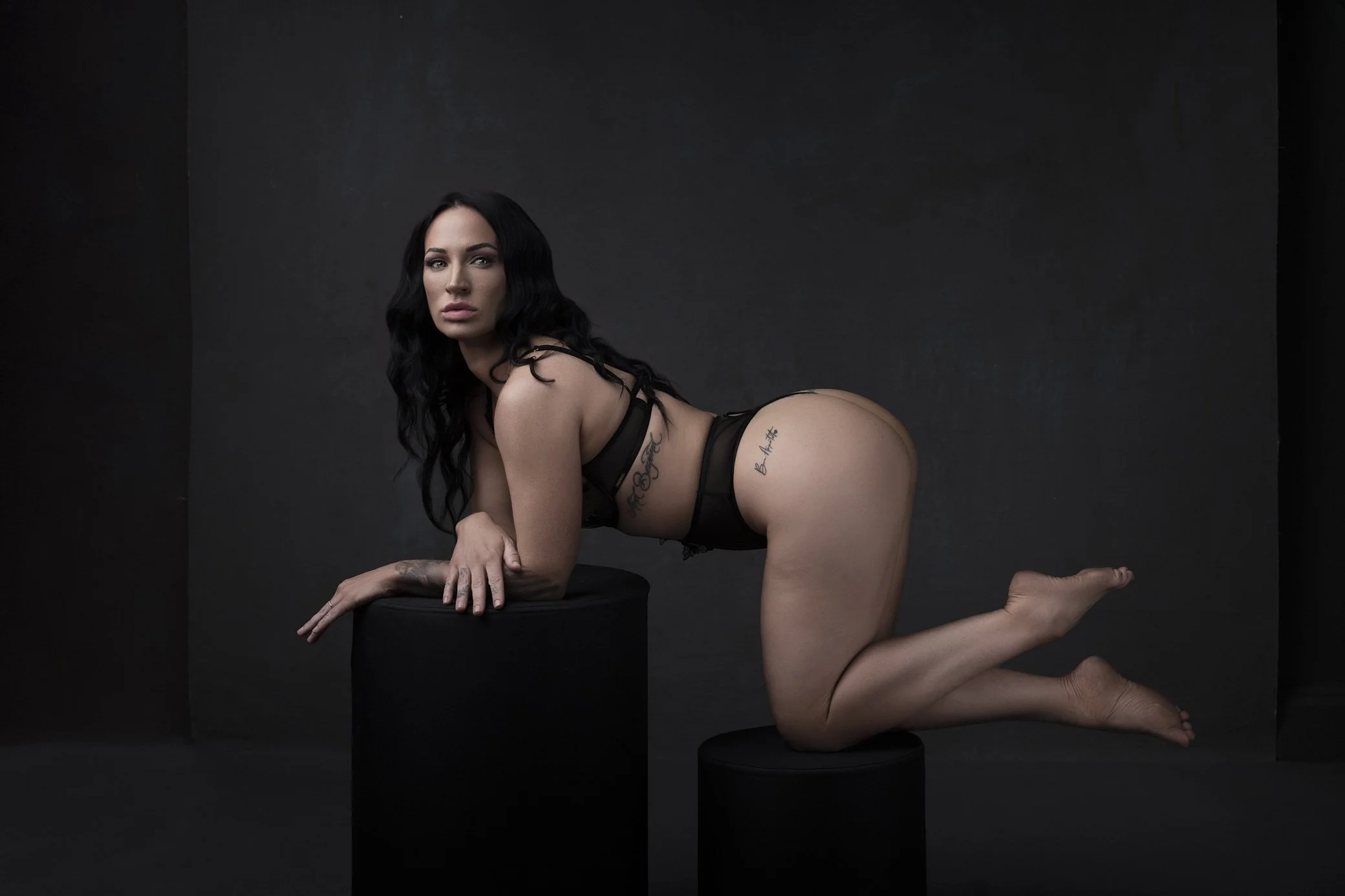 A woman with long black hair and tattoos, wearing black lingerie, is kneeling on one knee on a black stool and leaning forward with her hands on a larger black stool against a dark background.