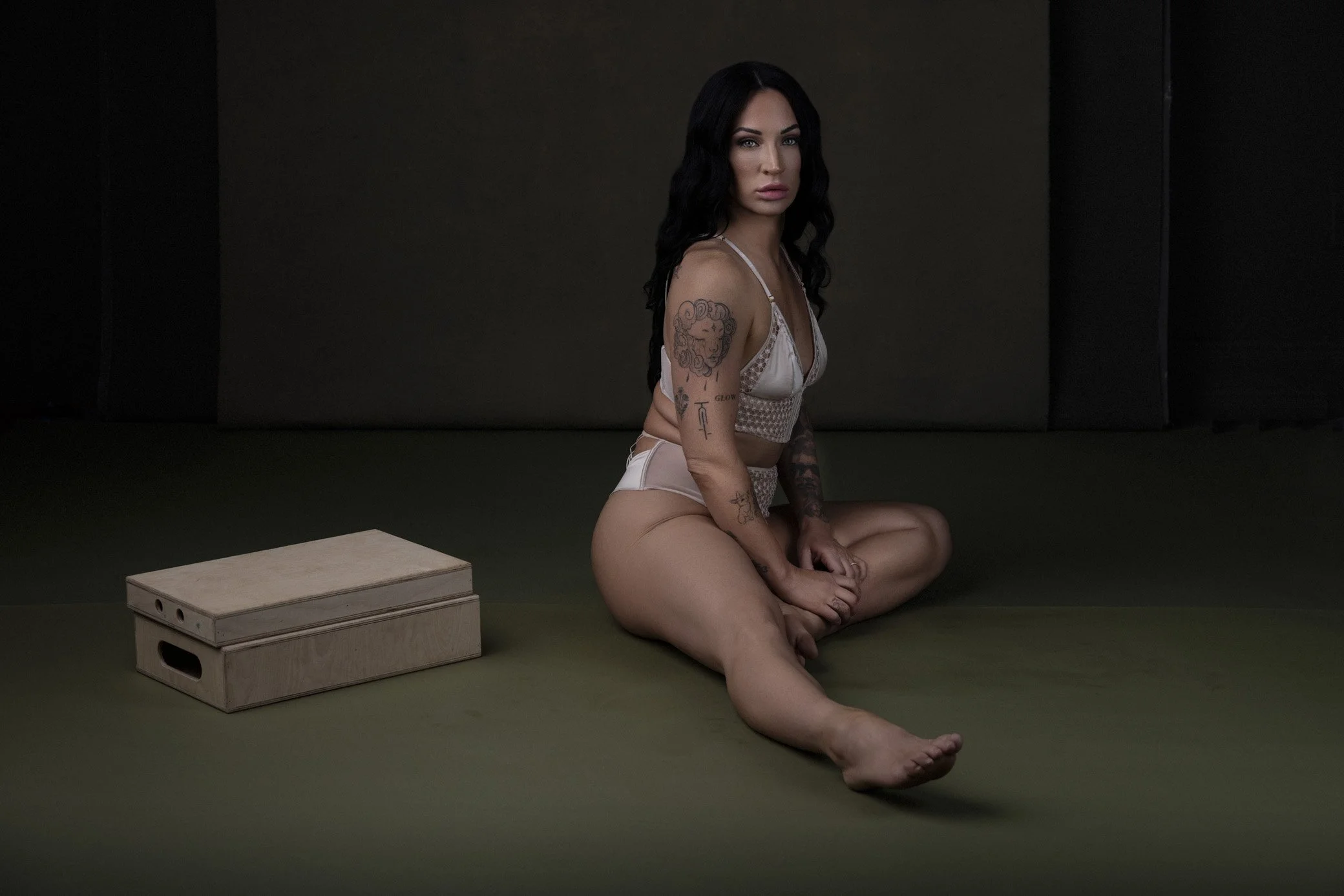 A woman with black hair and tattoos sitting cross-legged on a dark floor, wearing lingerie, next to a wooden box.